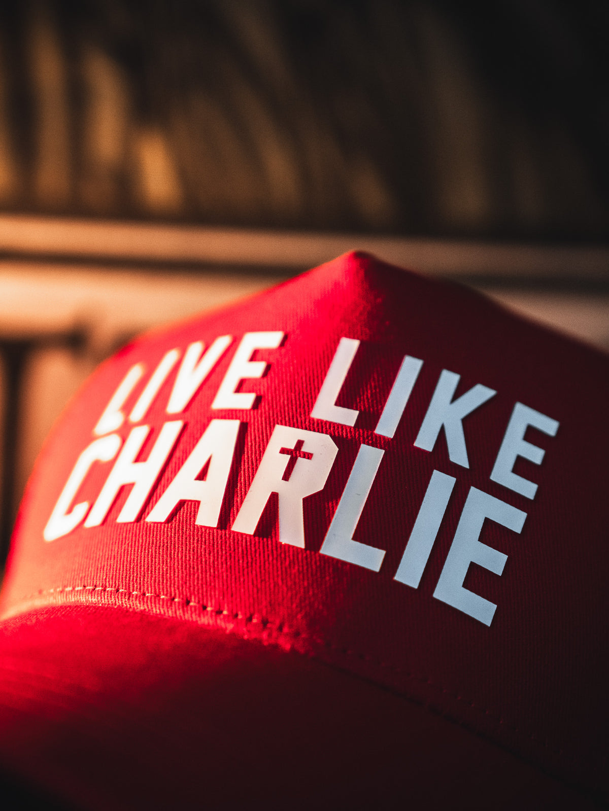 A close-up of the Born United Private Label Live Like Charlie Trucker Snapback, a red 5-panel curved brim hat featuring bold white "LIVE LIKE CHARLIE" lettering with the "I" in CHARLIE designed as a cross.