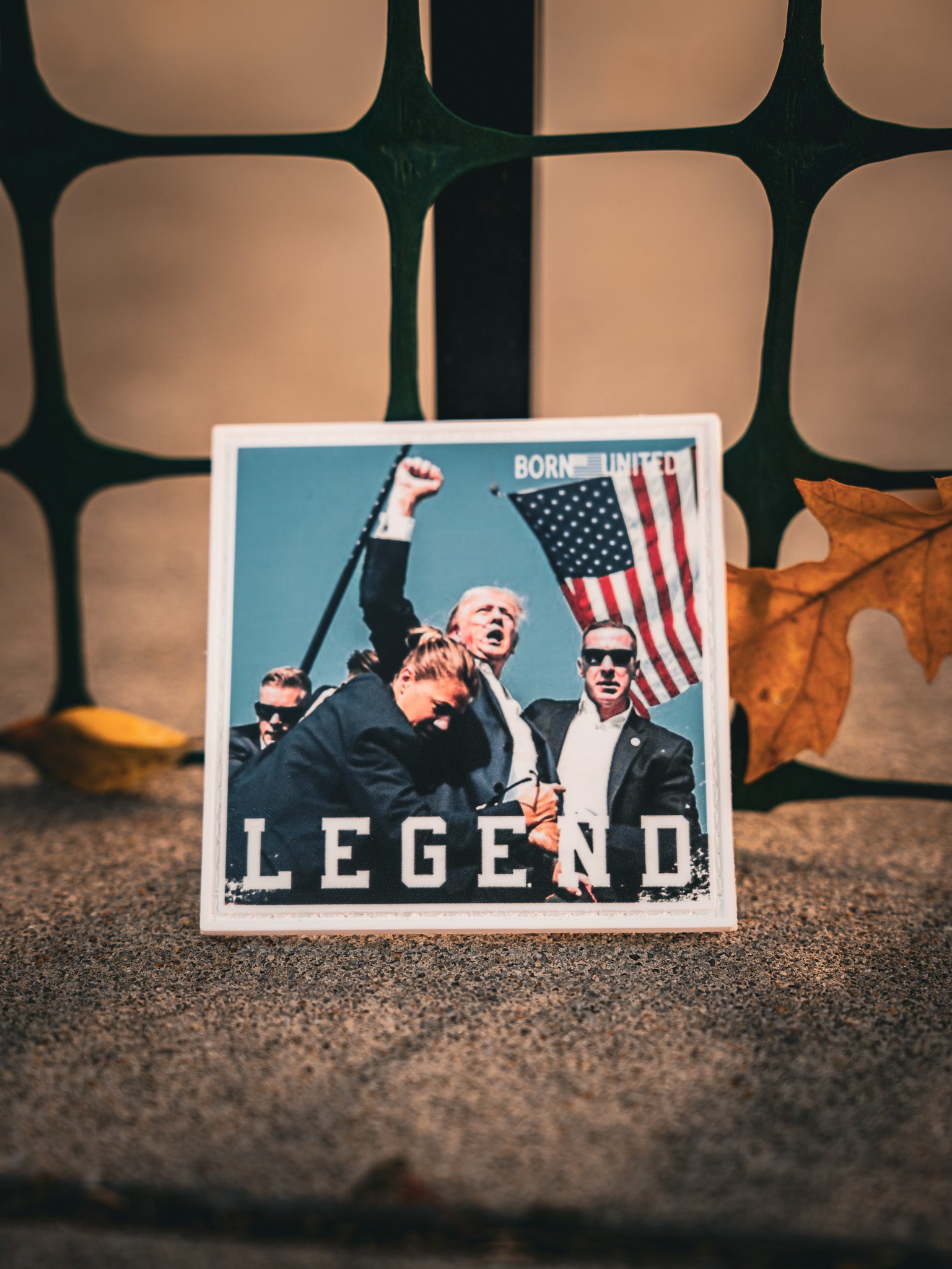 A yellow leaf lies near a card featuring the Forever Legend - Trump PVC Patch by Born United Private Label, displaying an image of a man with an American flag and bodyguards. The word LEGEND is printed on it, against a backdrop of a metal fence.