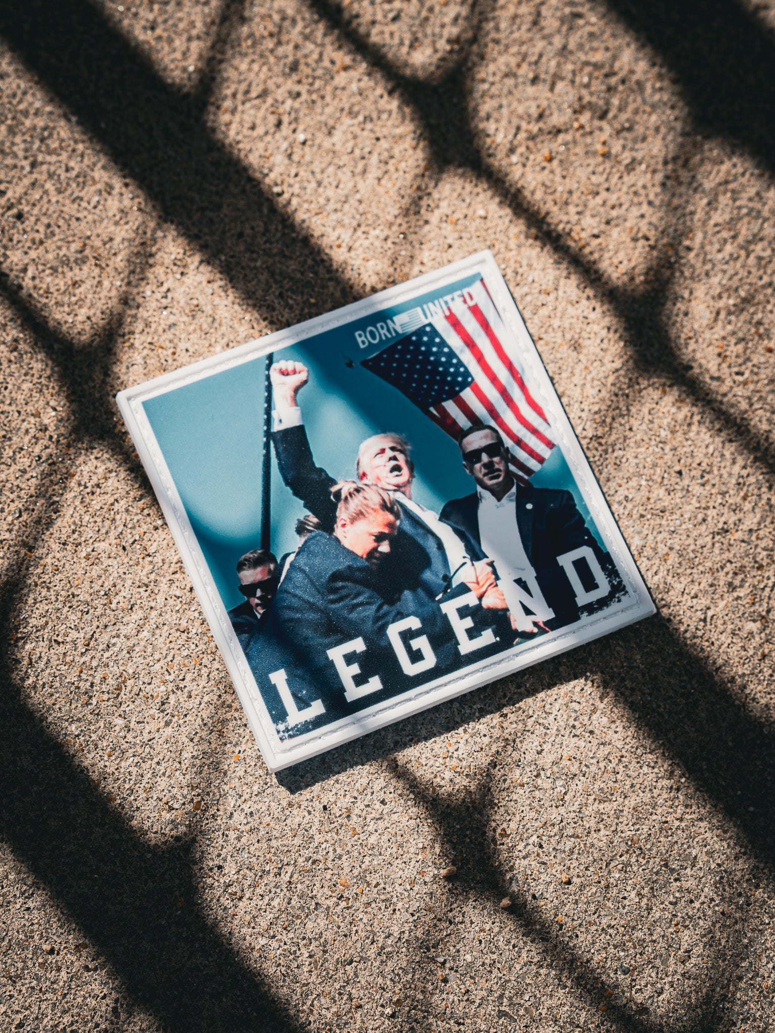 A square ground picture shows cheering people with one holding the American flag, marked LEGEND at the bottom. Shadows of a grid fall over it. This resembles the Forever Legend - Trump PVC Patch by Born United Private Label.