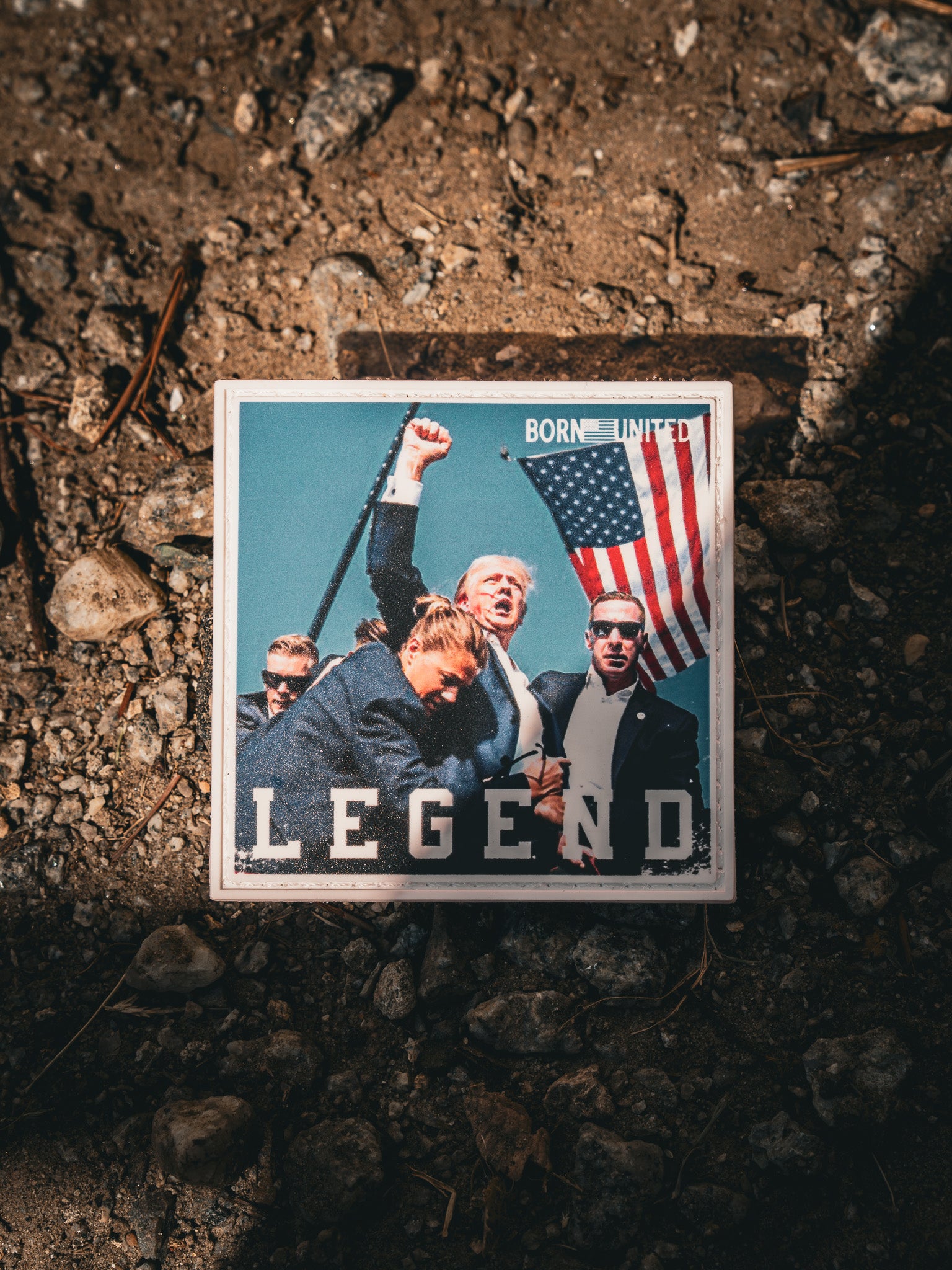 A square coaster has a stylized image of a suited man with a raised fist, surrounded by others, with an American flag visible. It features the text BORN UNITED and Forever Legend - Trump PVC Patch by Born United Private Label.