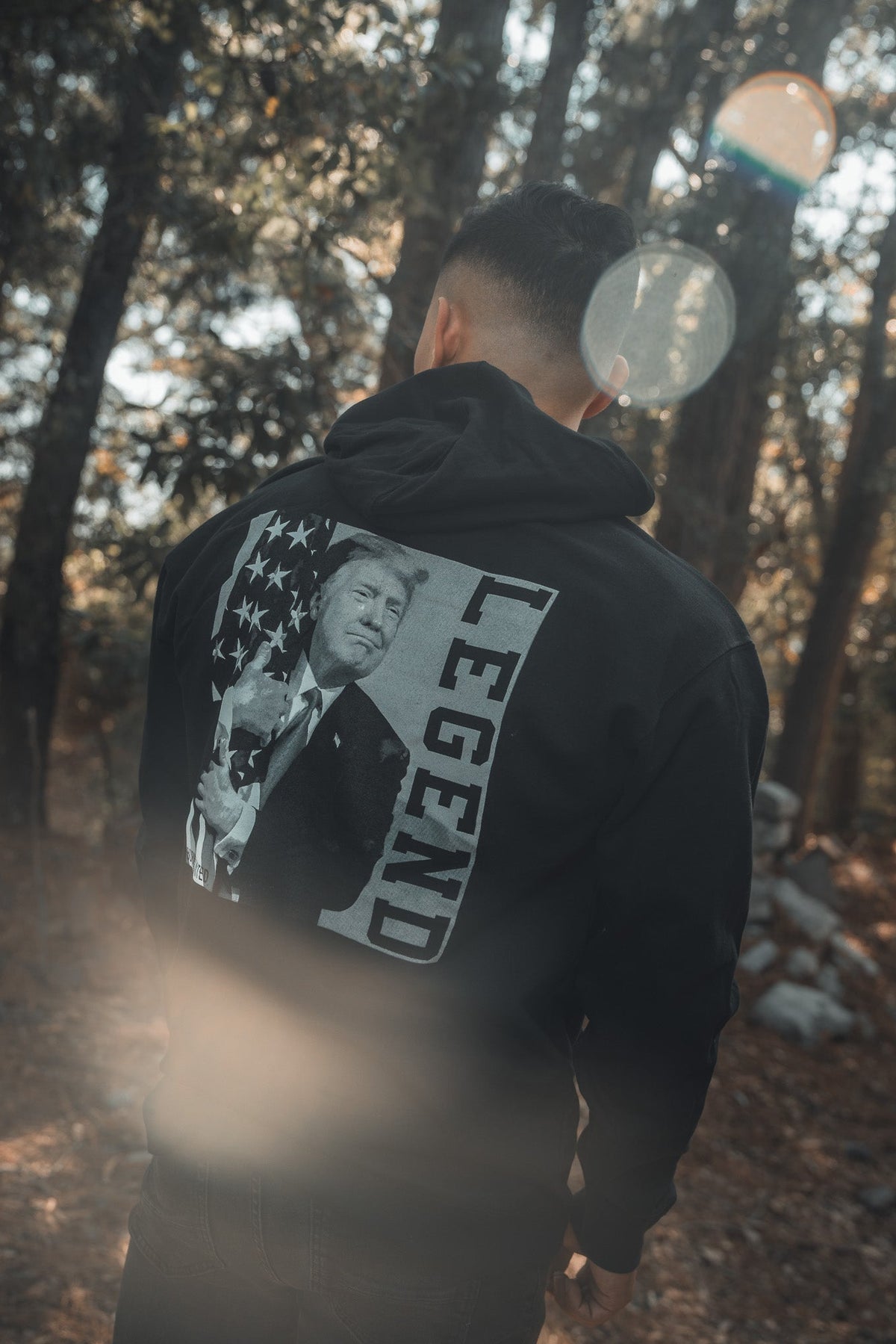 In a sunlit forest, a person wears the Born United Private Label "Legend - Trump Hoodie," a black sweatshirt featuring an image of Trump holding an American flag with the word LEGEND beside him.