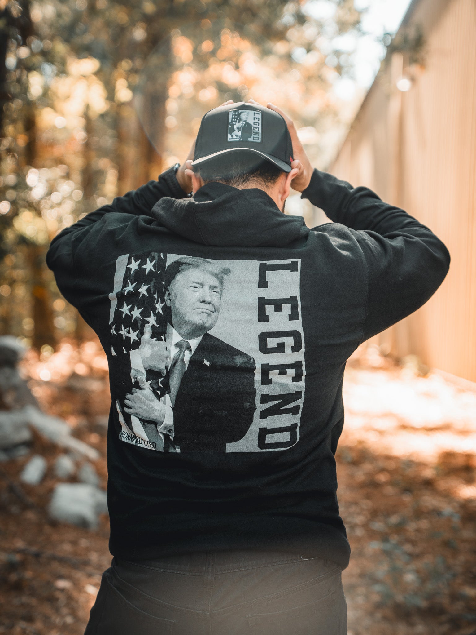 Outdoors, a person wears the Born United Private Label Legend Trump Hoodie and cap. The hoodie’s back shows Donald Trump holding a cat by an American flag, with LEGEND printed vertically.