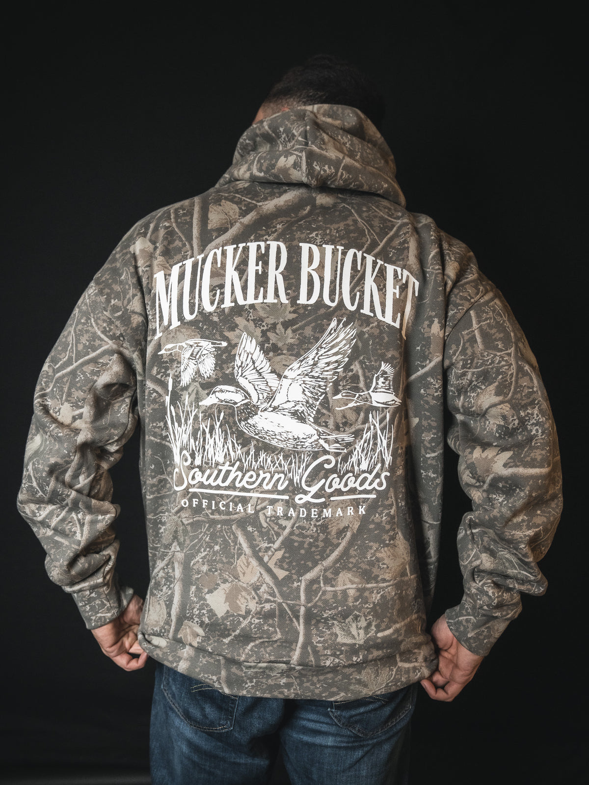 A person wearing the Mallard Deadwood Camo Hoodie by Mucker Bucket, featuring flying ducks and white Southern Goods Official Trademark print on the back, stands against a black background.