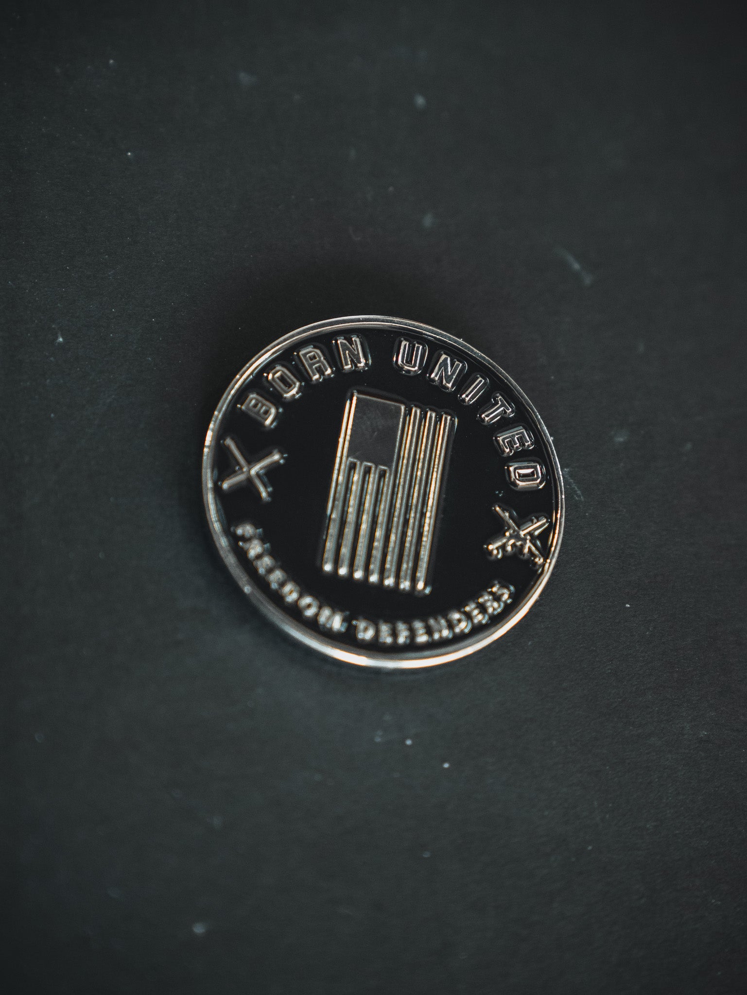 A round, metallic pin on a dark surface features an American flag at the center with Born United and Freedom Defenders text. Two cross motifs symbolize unity for a brighter future. Product: Saints and Sinners Challenge Coin by Born United Private Label.