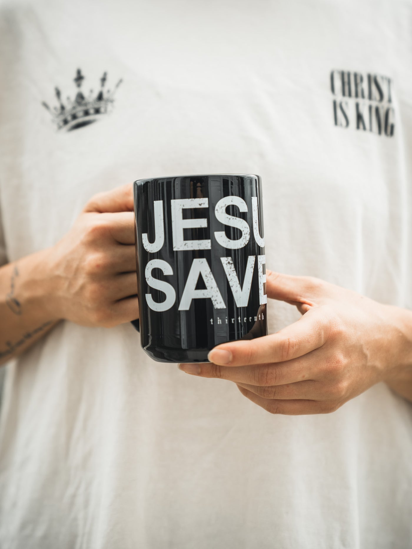 A person holds the 13th's Jesus Saves Mug with bold white lettering, wearing a white CHRIST IS KING shirt featuring a striking crown graphic.