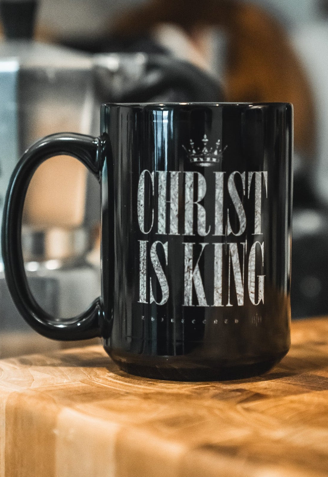 The Christ is King Mug by 13th, featuring bold white CHRIST IS KING lettering on black, sits on a wooden countertop with a blurred silver stovetop espresso maker in the background, expressing Christian faith.