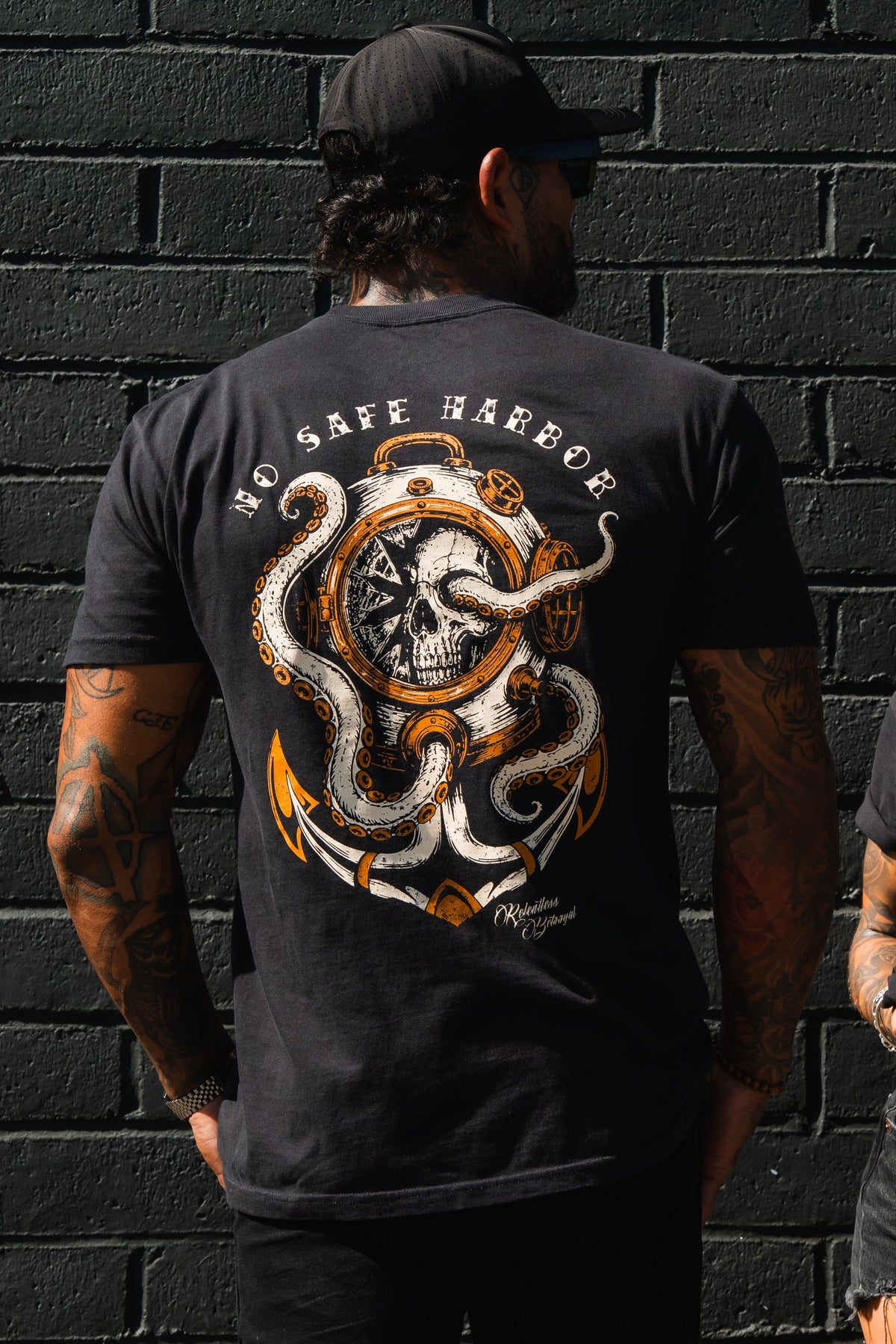 A man stands against a dark brick wall, wearing the Relentless Betrayal No Safe Harbor Premium T-Shirt featuring a skull, compass, and octopus nautical graphic.