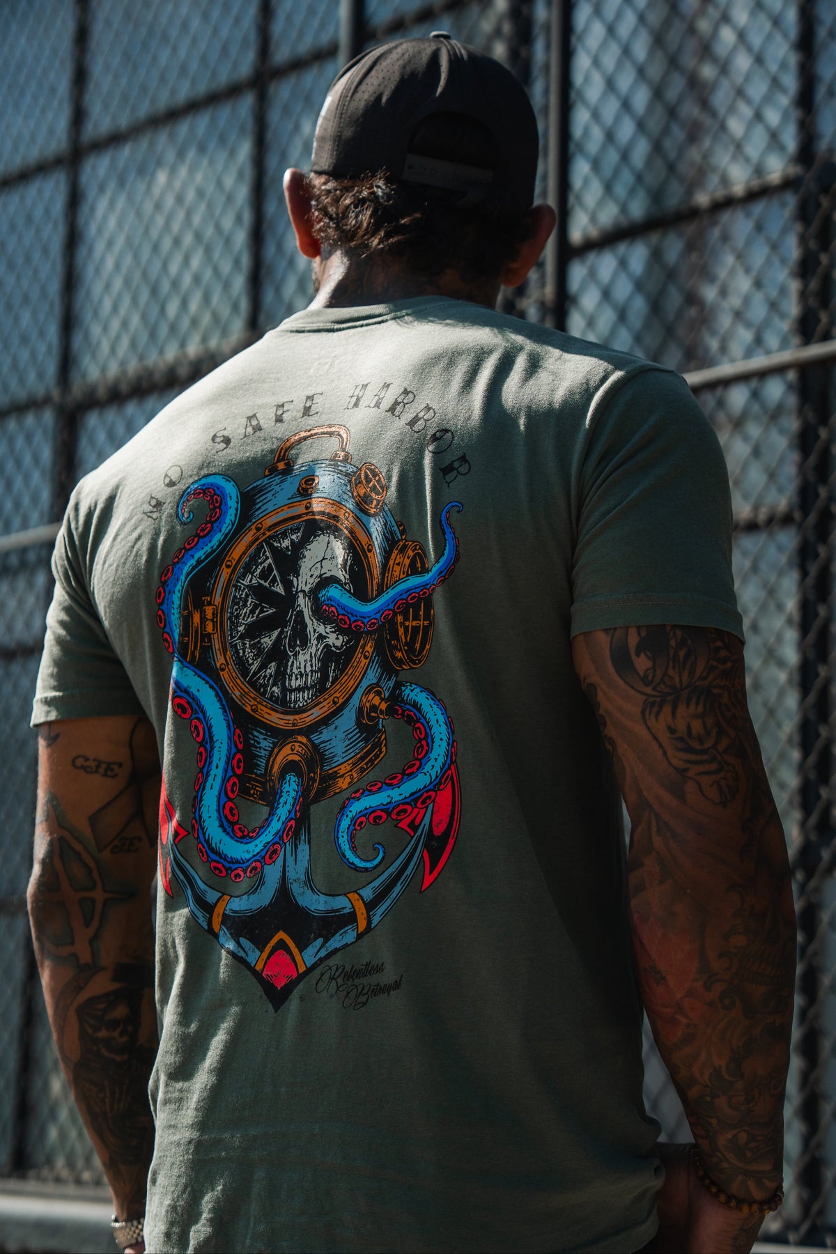 A tattooed man in a cap wears the No Safe Harbor Premium T-Shirt - Olive by Relentless Betrayal—an army-green unisex tee with a nautical skull and octopus design.
