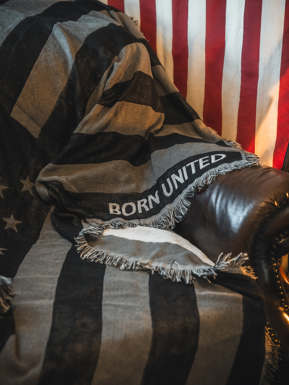 A leather chair is draped with the Born United Private Label Blackout American Flag Jacquard Blanket, featuring a black-and-white flag design and "BORN UNITED" text, set before a red and white striped hanging American flag.