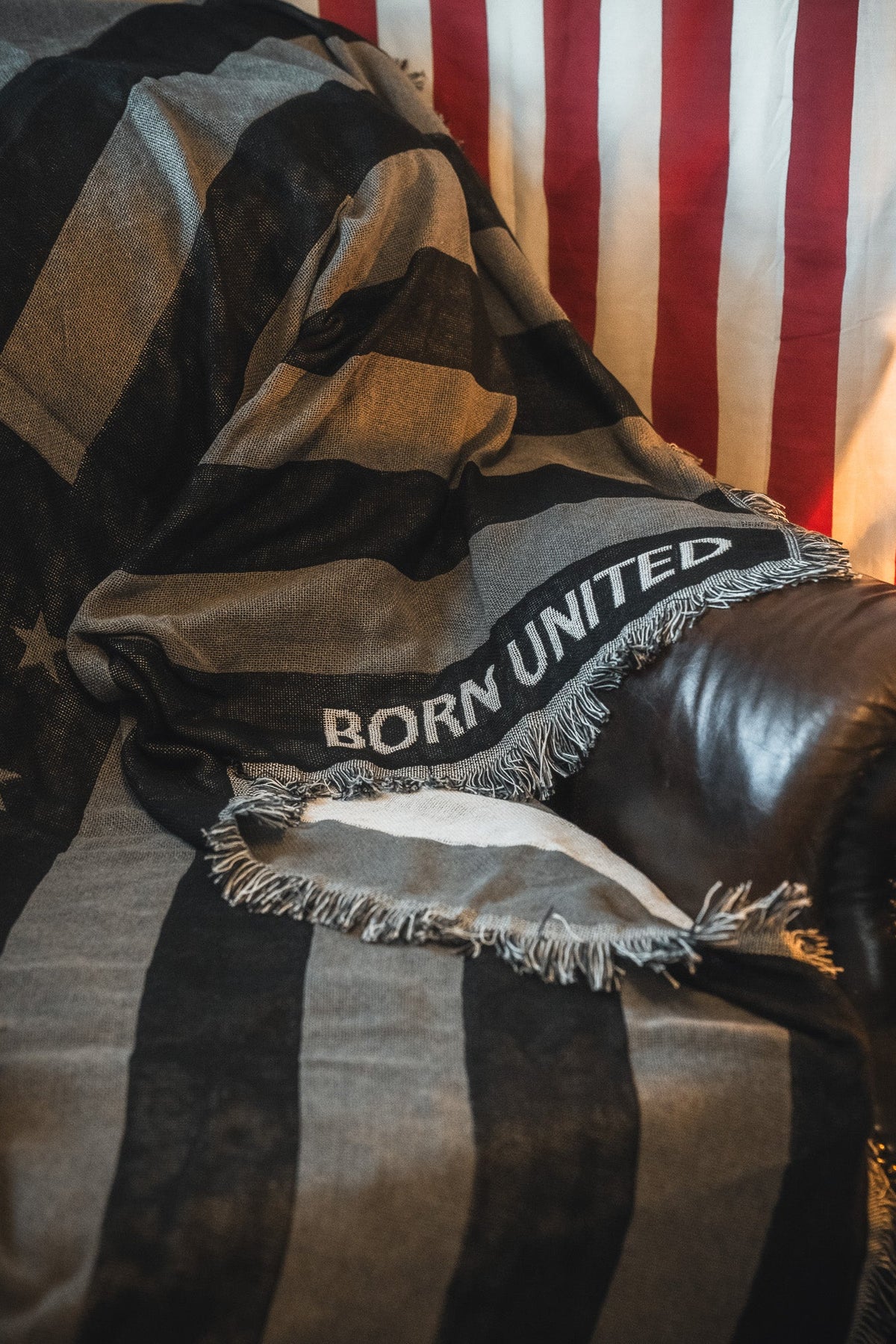 A leather chair is draped with the Born United Private Label Blackout American Flag Jacquard Blanket, featuring a black-and-white flag design and "BORN UNITED" text, set before a red and white striped hanging American flag.