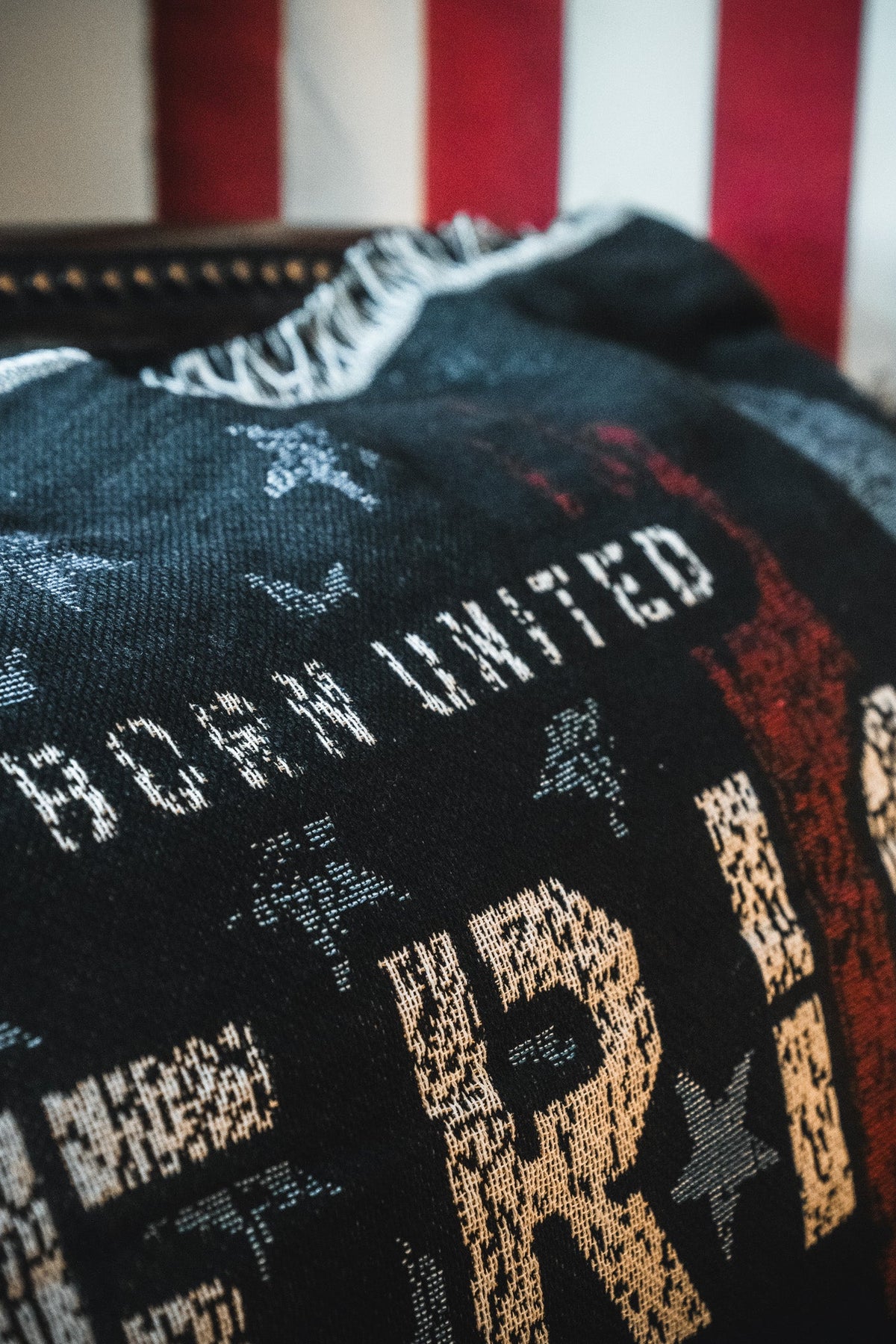 A close-up of the American Heroes Jacquard Blanket by Born United Private Label, showcasing woven stars and stripes in red, white, and blue with the phrase "BORN UNITED." A blurred American flag appears in the background.