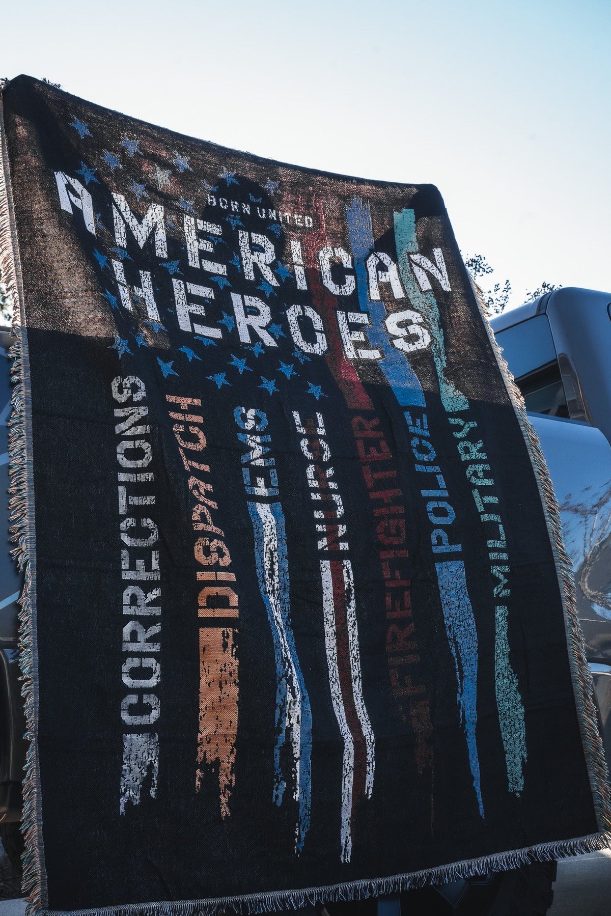 The Born United Private Label American Heroes Jacquard Blanket displays “American Heroes” with colorful faded stripes labeled Corrections, Dispatch, EMS, Firefighter, Police, and Military—perfect for draping over a vehicle.