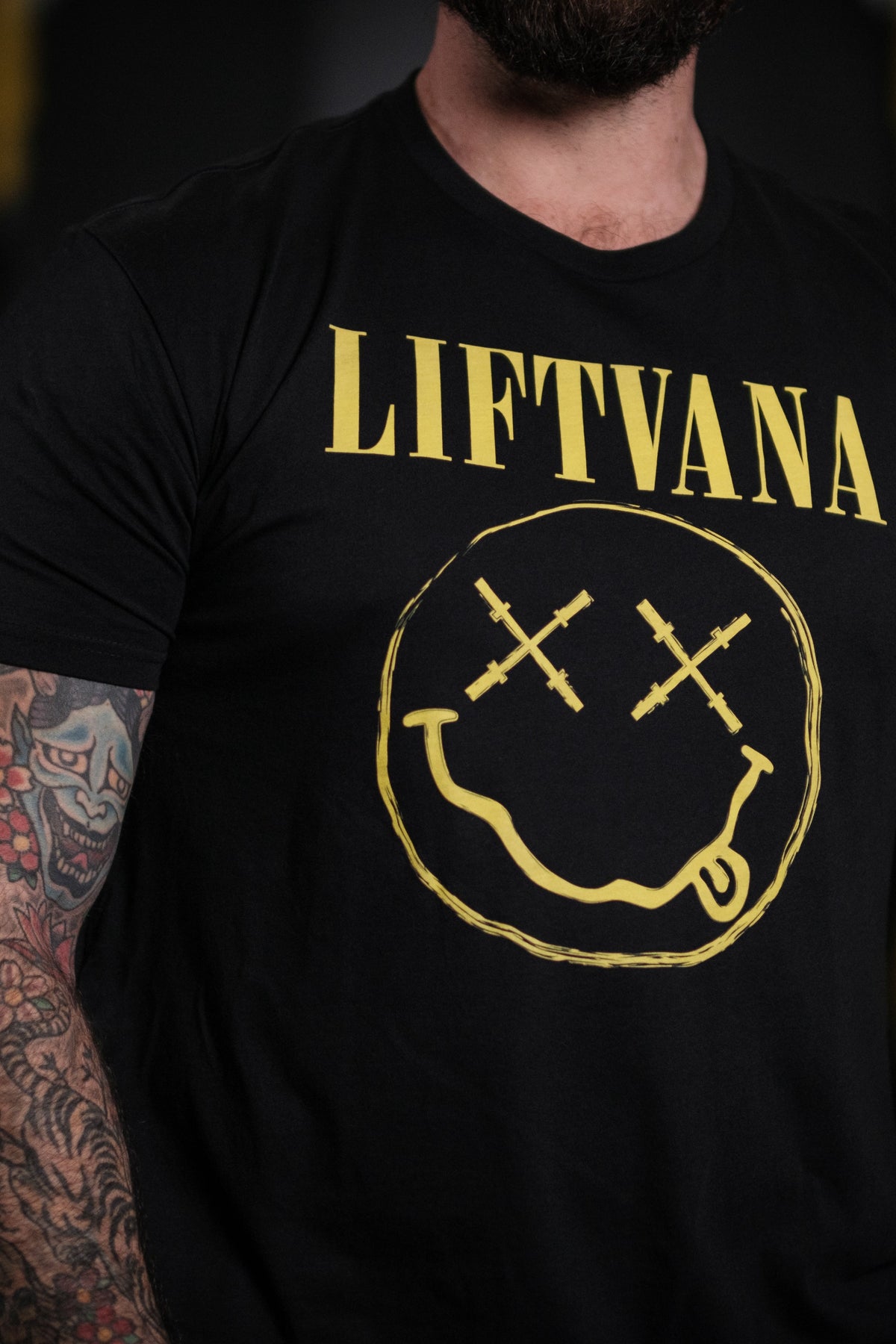 A muscular man with tattooed arms wears the Liftvana Mens Tee by Tango Charlie Apparel, featuring Lift•Va•Na text and a smiley face with barbell eyes—grunge vibes inspired by the iconic Nirvana logo.