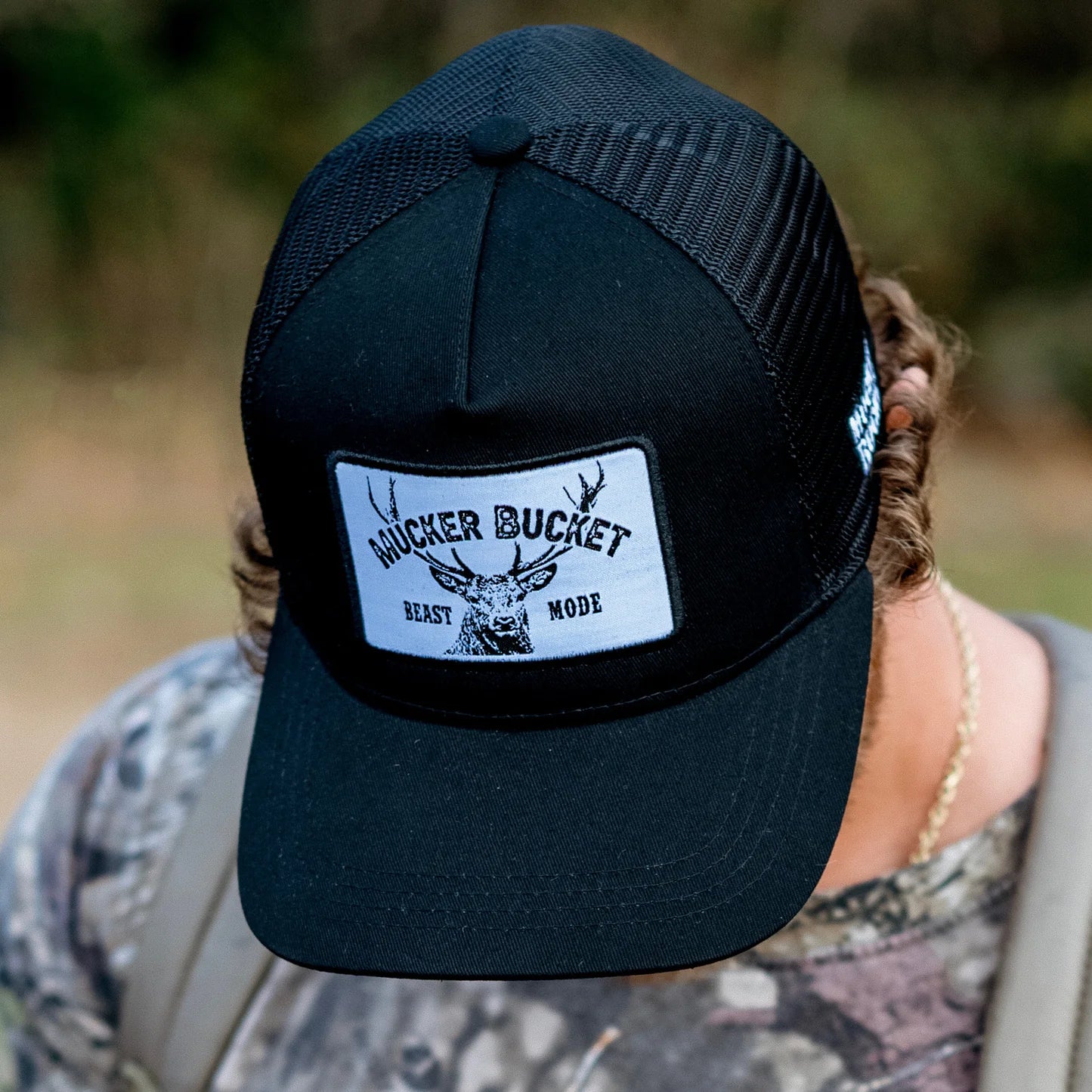 Wearing Mucker Buckets Beast Mode OG Trucker Bucket snapback, featuring a deer head design, and dressed in camo, they stand against a blurred green backdrop—ideal for any dedicated hunter or white tail deer enthusiast.
