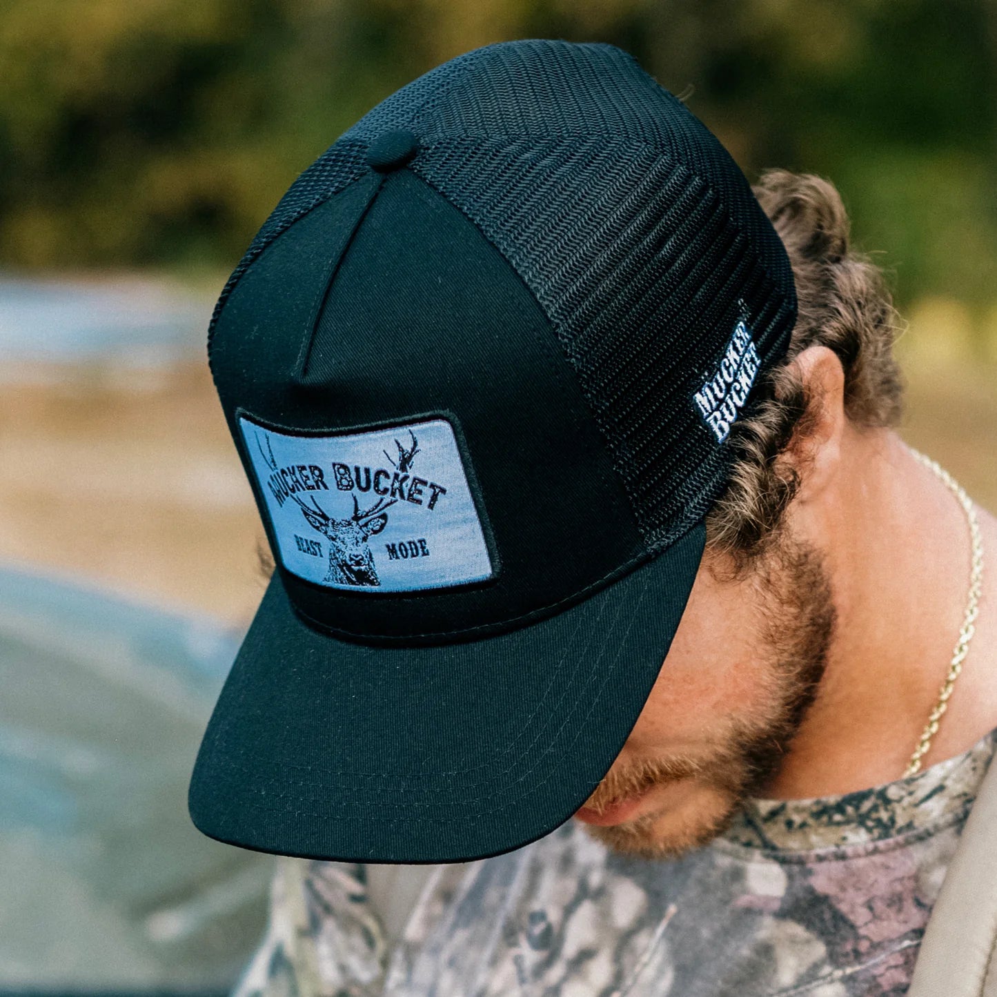 A man in a camouflage shirt and a black Beast Mode OG Trucker Bucket hat by Mucker Bucket stands against blurred greenery. The hats mesh back enhances its rugged style, ideal for outdoor adventures.