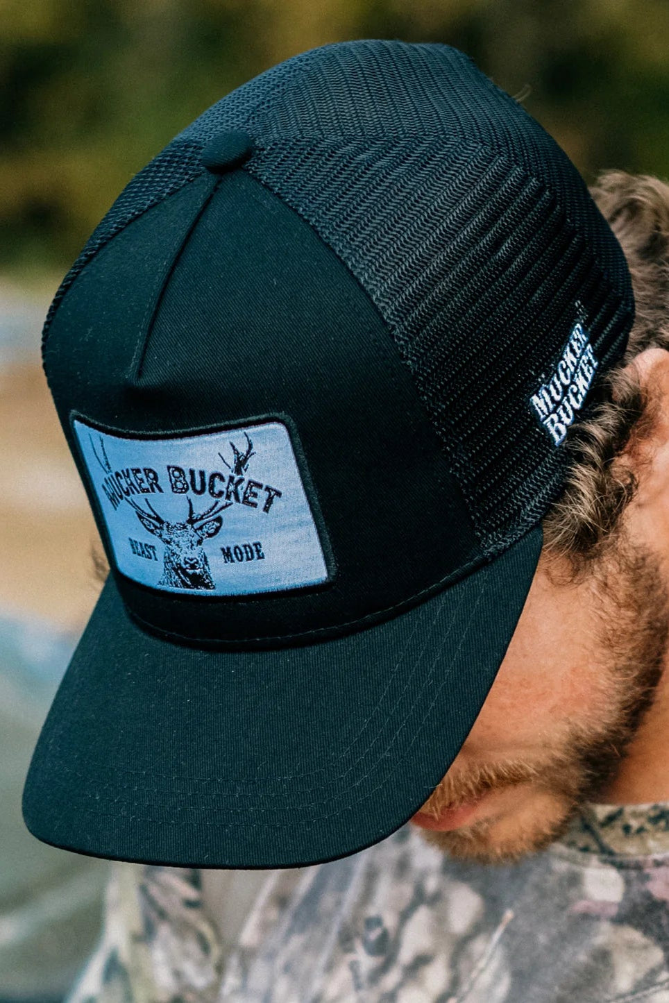 A man in a camouflage shirt and a black Beast Mode OG Trucker Bucket hat by Mucker Bucket stands against blurred greenery. The hats mesh back enhances its rugged style, ideal for outdoor adventures.