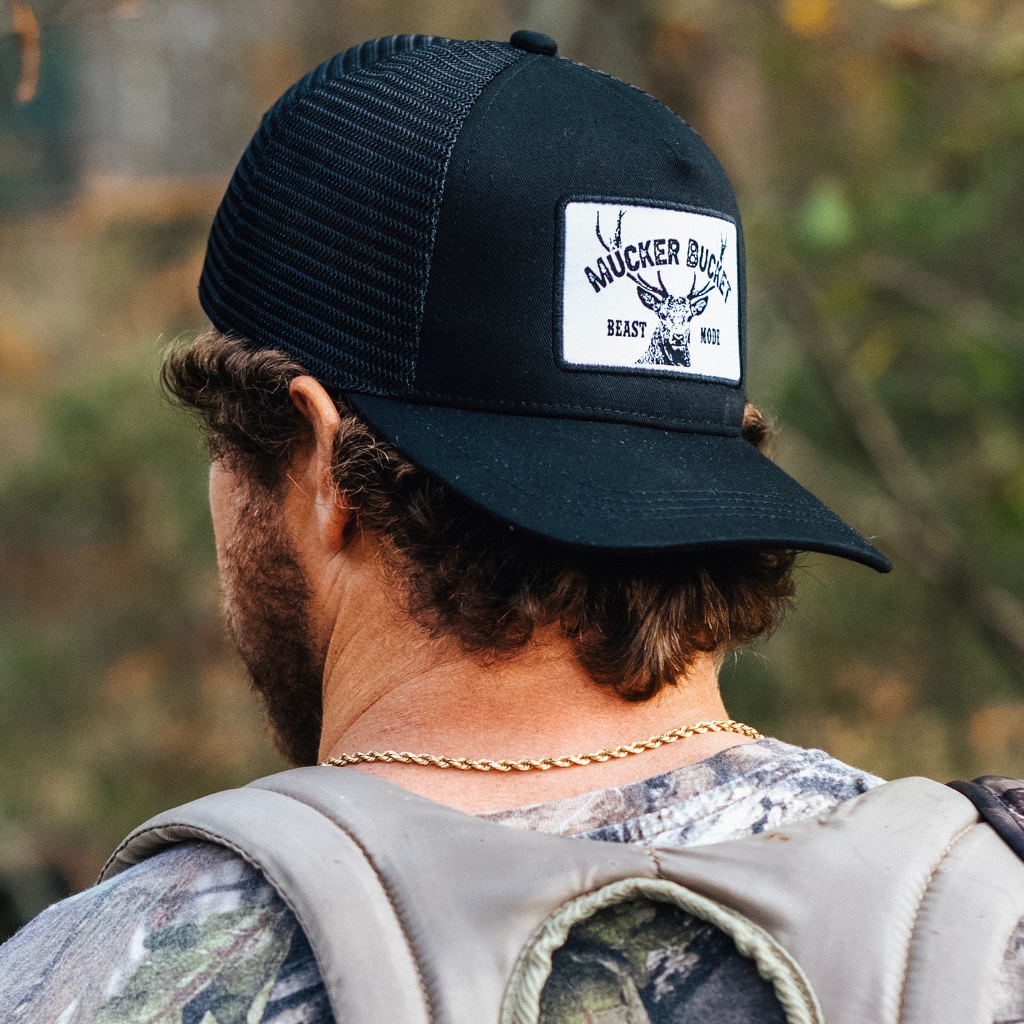 A person with curly hair wears a black mesh back trucker snapback hat from Mucker Bucket, featuring the Beast Mode patch with antler design. Theyre dressed in camo clothing and a gold chain, standing outdoors.