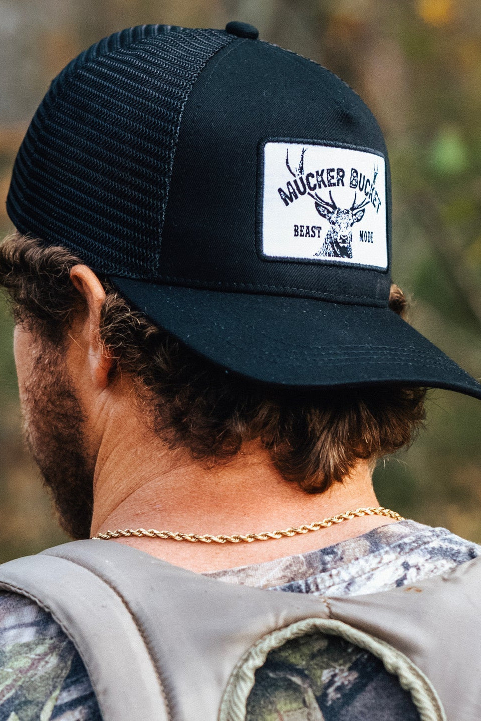 A person with curly hair wears a black mesh back trucker snapback hat from Mucker Bucket, featuring the Beast Mode patch with antler design. Theyre dressed in camo clothing and a gold chain, standing outdoors.
