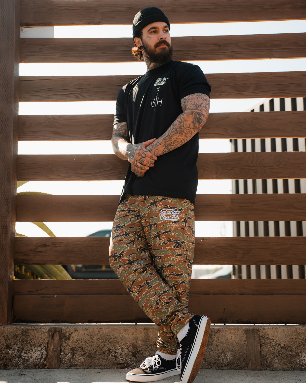 A tattooed man in a black beanie and t-shirt stands by a wooden fence, sporting Tango Charlie Apparel’s Men’s Classic Premium Joggers | Snipes & Stripes, camo print with quick-dry fabric, paired with black sneakers.