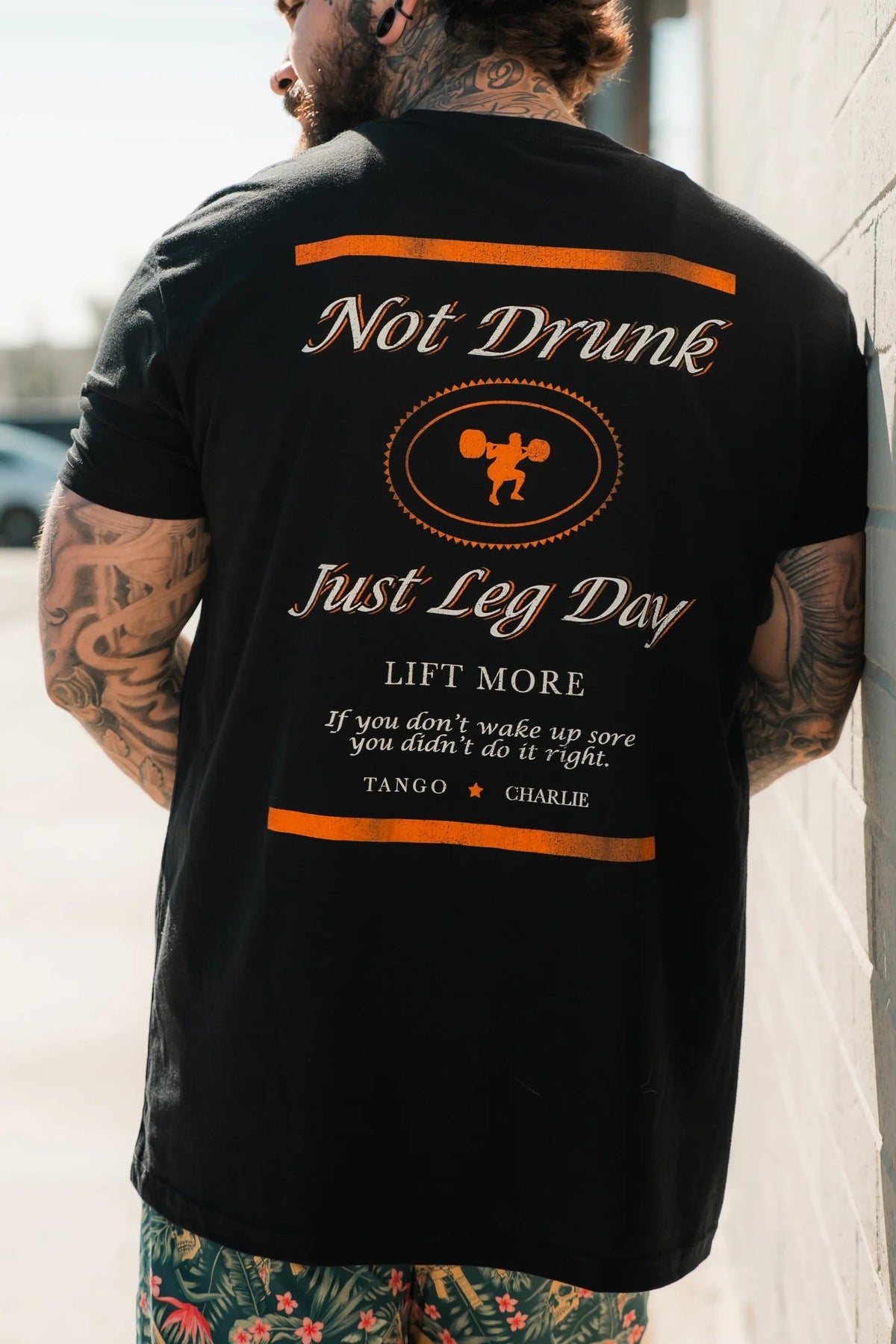A tattooed person faces away, wearing the Just Leg Day - Black workout tee by Tango Charlie Apparel. The shirt features bold orange and white text: “Not Drunk, Just Leg Day. Lift More. If you don’t wake up sore, you didn’t do it right.”.