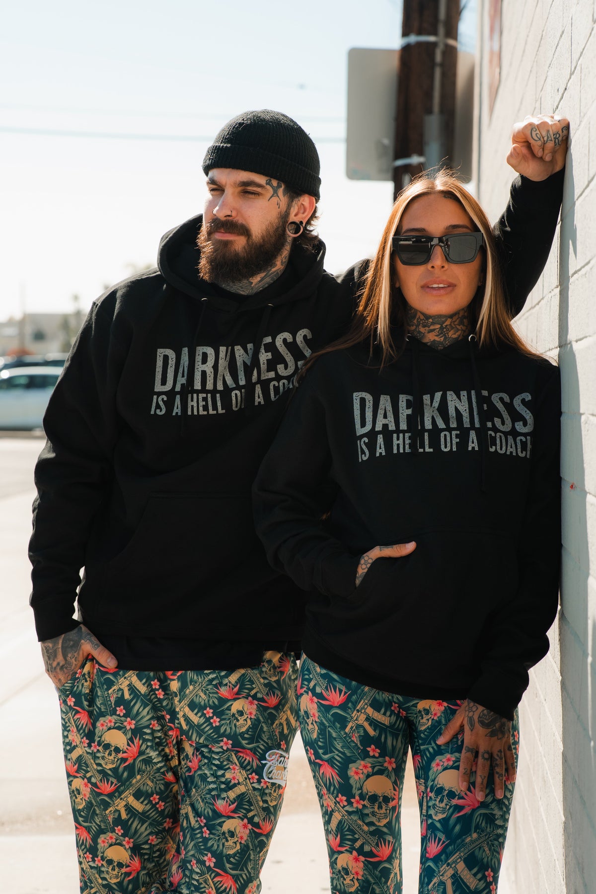 A man and woman stand outside in matching Tango Charlie Apparel "Darkness is a Hell of a Coach" hoodies and colorful skull-patterned pants, embodying victory over darkness in bold gymwear.