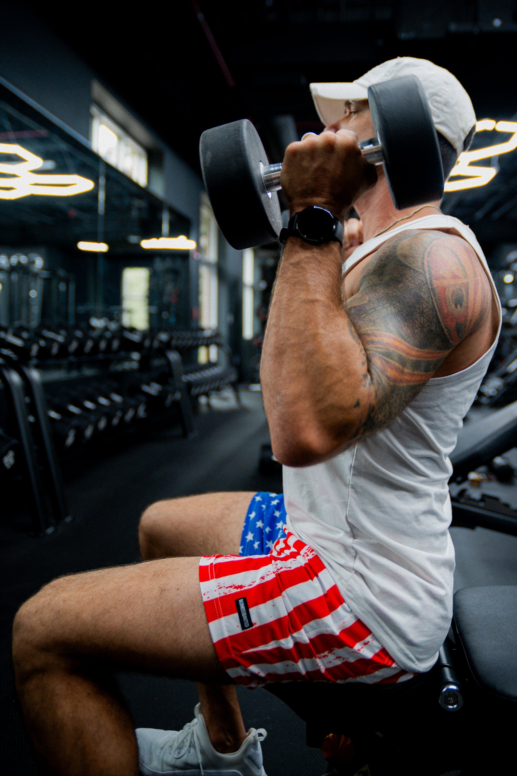 A man in a white tank top, cap, and Born United Private Label Mens Performance Hybrid Shorts 5.5 | Stars & Stripes lifts a dumbbell on a gym bench, displaying his tattooed arm amid gym equipment and mirrors.