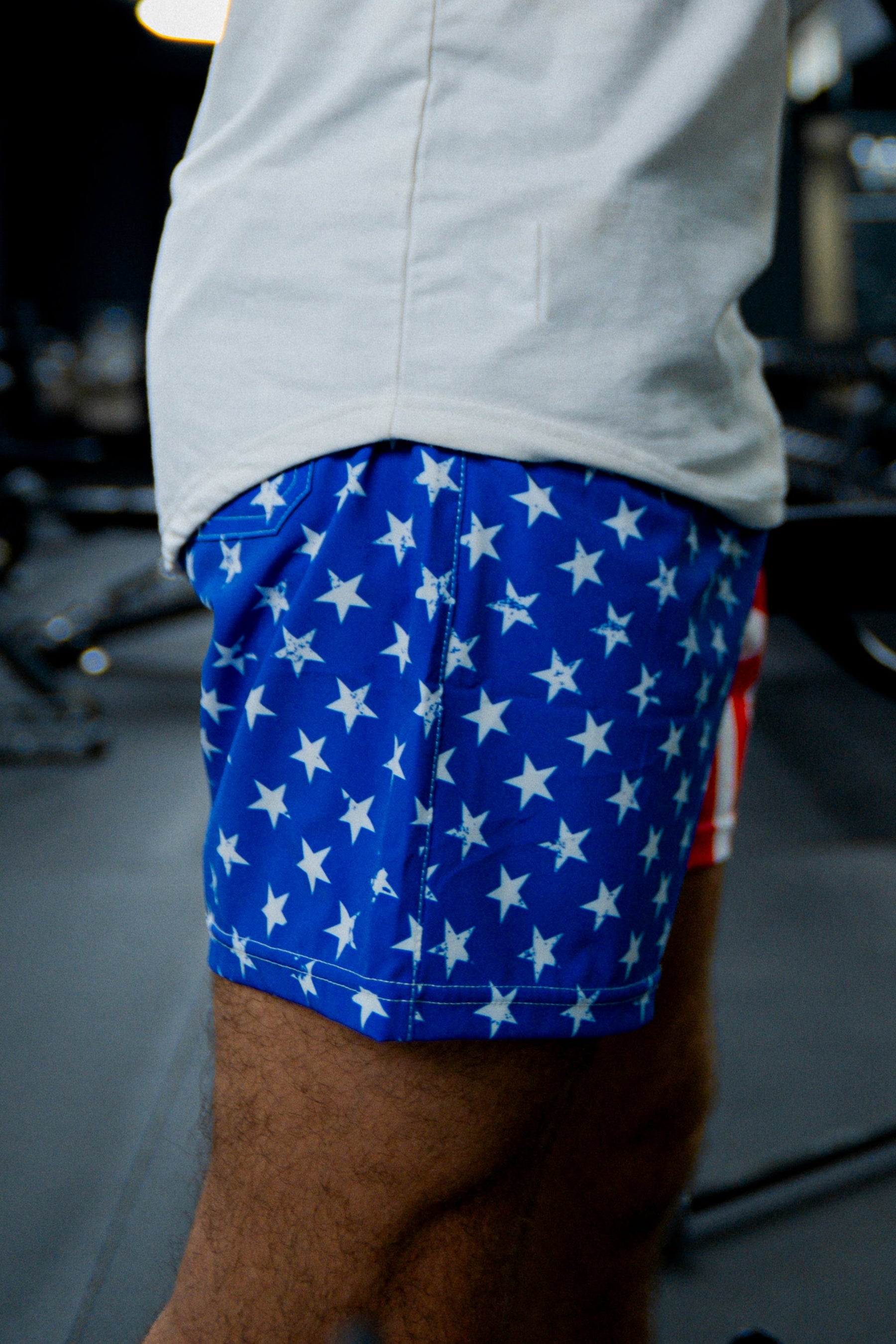 A person wears Born United Private Label Mens Performance Hybrid Shorts 5.5 | Stars & Stripes, featuring a blue fabric with white stars. Only their lower torso and upper legs are visible, with blurred gym equipment in the background.
