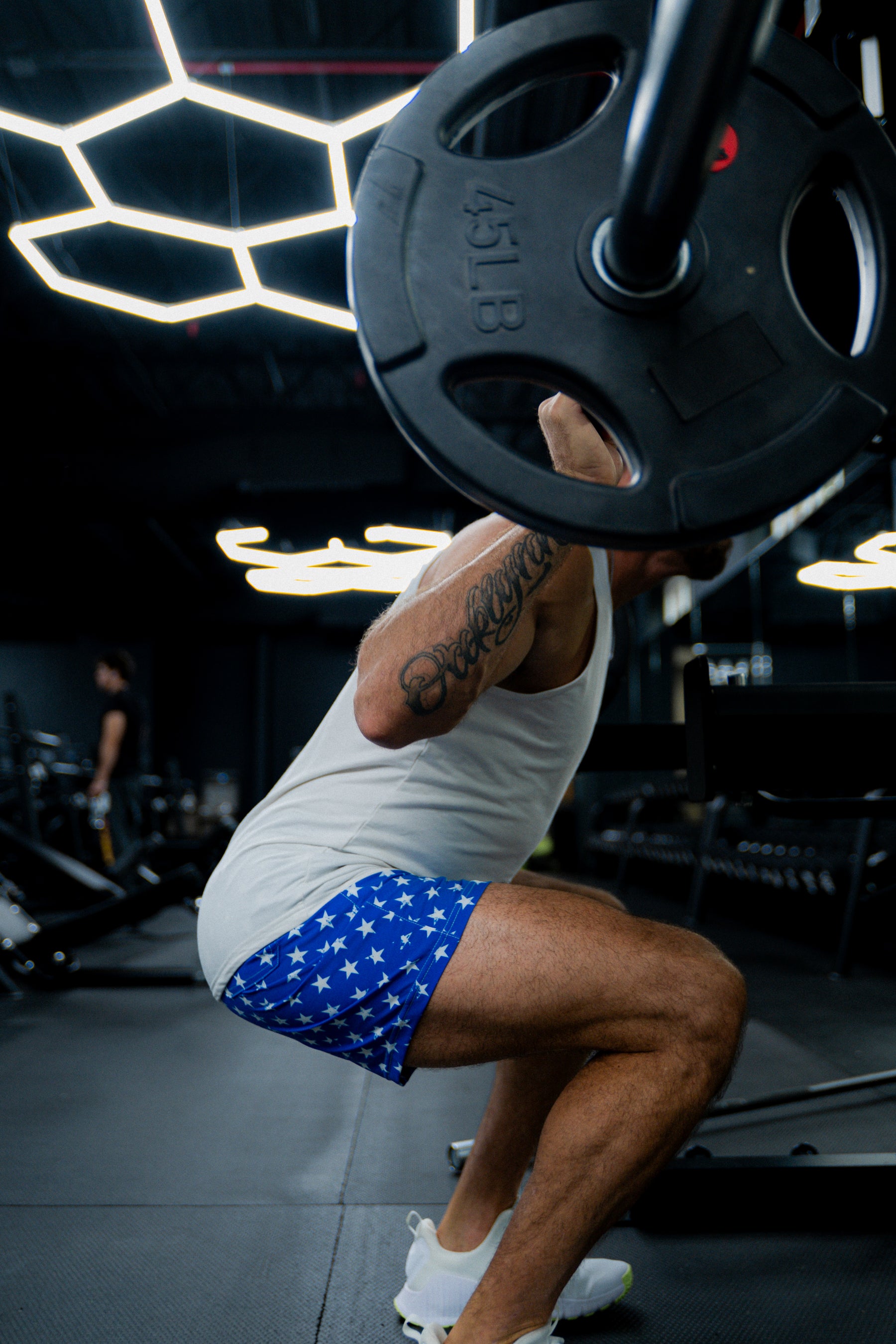 A man squats in the gym wearing a white tank top, white sneakers, and Born United Private Label Mens Performance Hybrid Shorts 5.5 | Stars & Stripes. Modern gym lighting and equipment appear in the background.