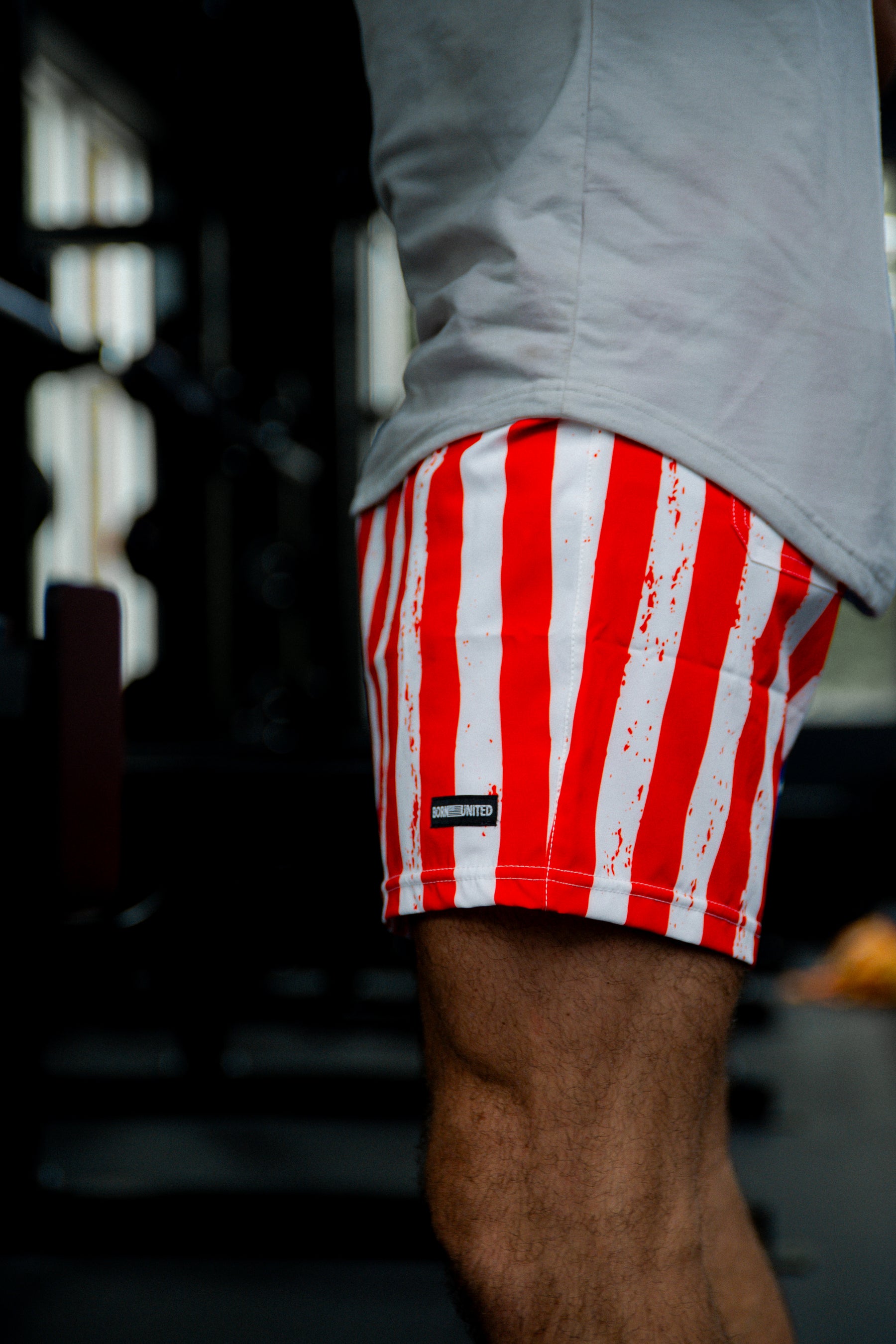 A person in Born United Private Label Mens Performance Hybrid Shorts 5.5 | Stars & Stripes and a white shirt stands indoors, with gym equipment blurred in the background.