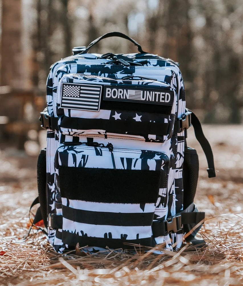 The Premium Tac-Pac Ultimate Bundle by Born United Private Label, featuring a black and white backpack with an American flag patch and BORN UNITED text, rests on dried pine needles amid a sunlit forest with blurred trees in the background.