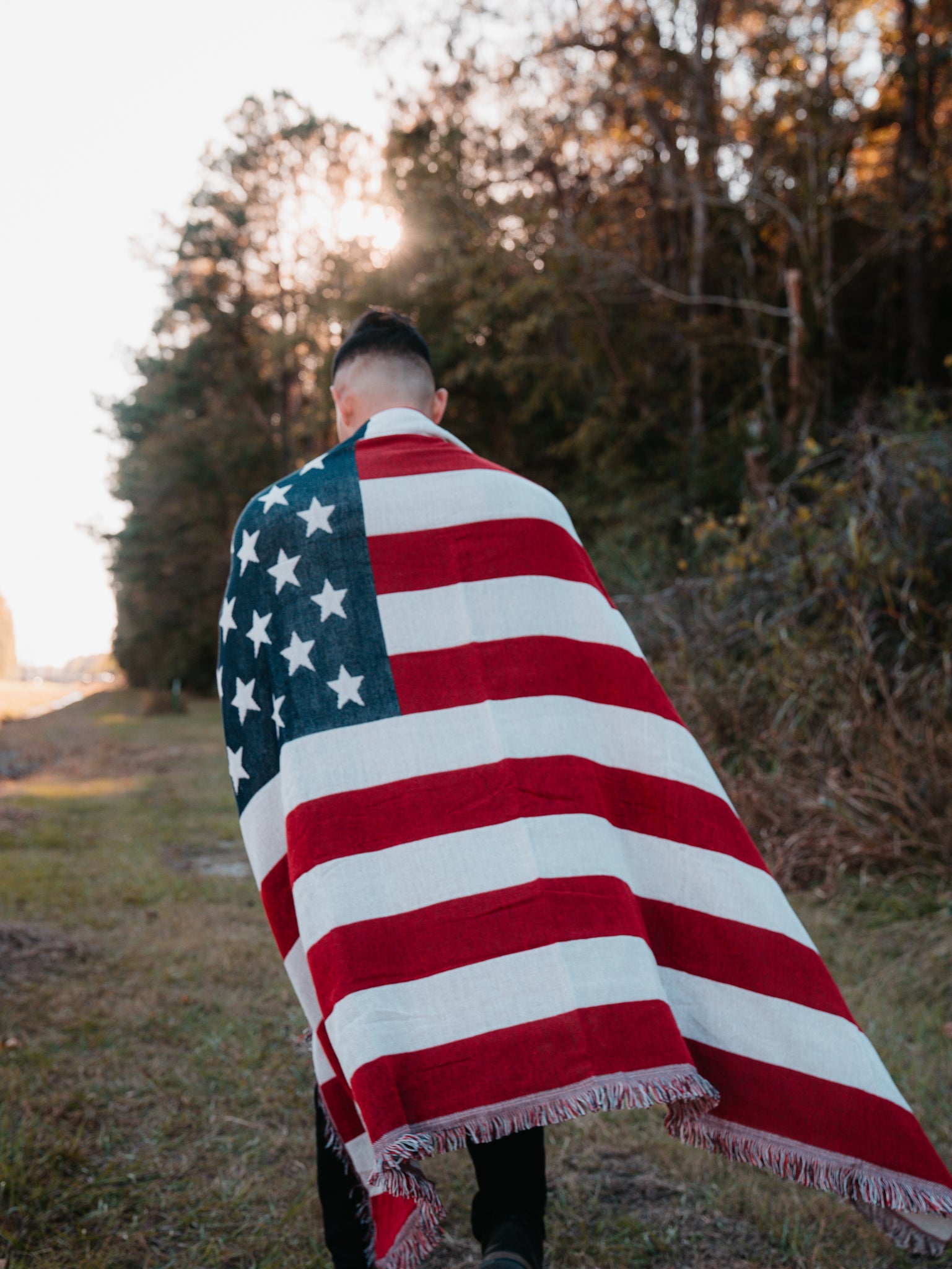 Wrapped in the American Flag Jacquard Blanket by Born United Private Label, a person strolls along a grassy path. The setting sun casts a warm light through the trees, creating a serene backdrop for the journey.