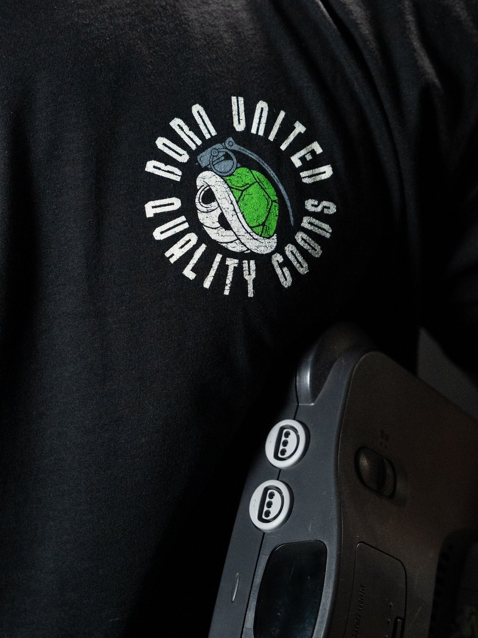 A man wearing a premium tee featuring a turtle graphic and the Born United Private Label logo holds a gray Super Merica Bros retro game console with two visible controller ports.