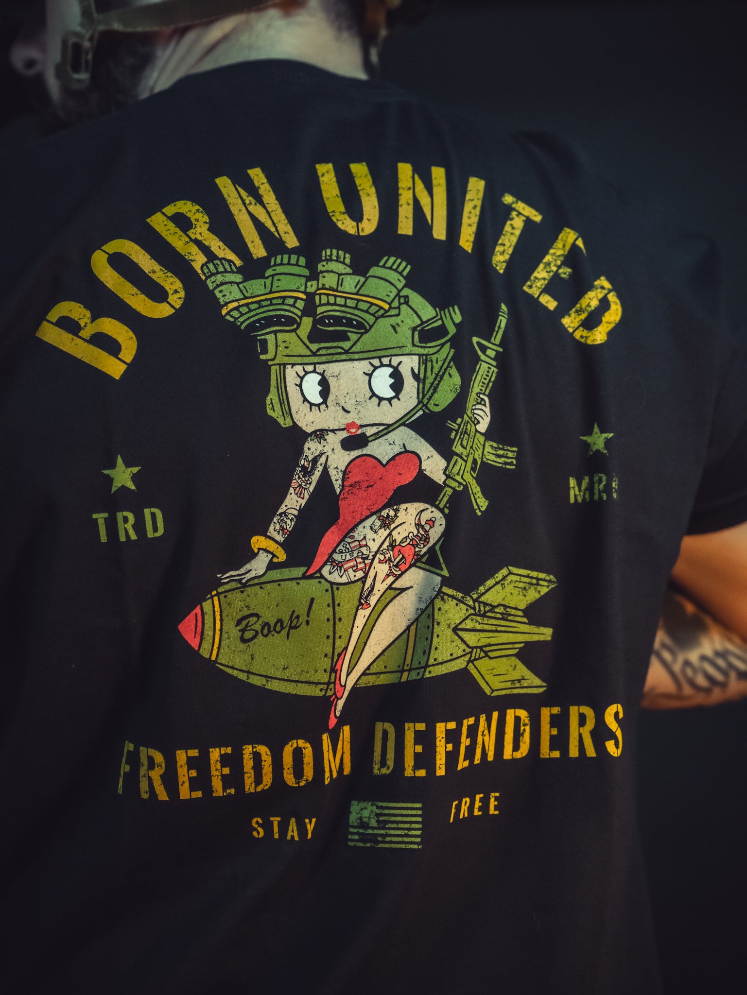A person wearing the Betty Boop Tee by Born United Private Label, featuring a colorful cartoon pin-up girl sitting on a bomb with BORN UNITED above and FREEDOM DEFENDERS below, surrounded by stars and elements celebrating defenders.