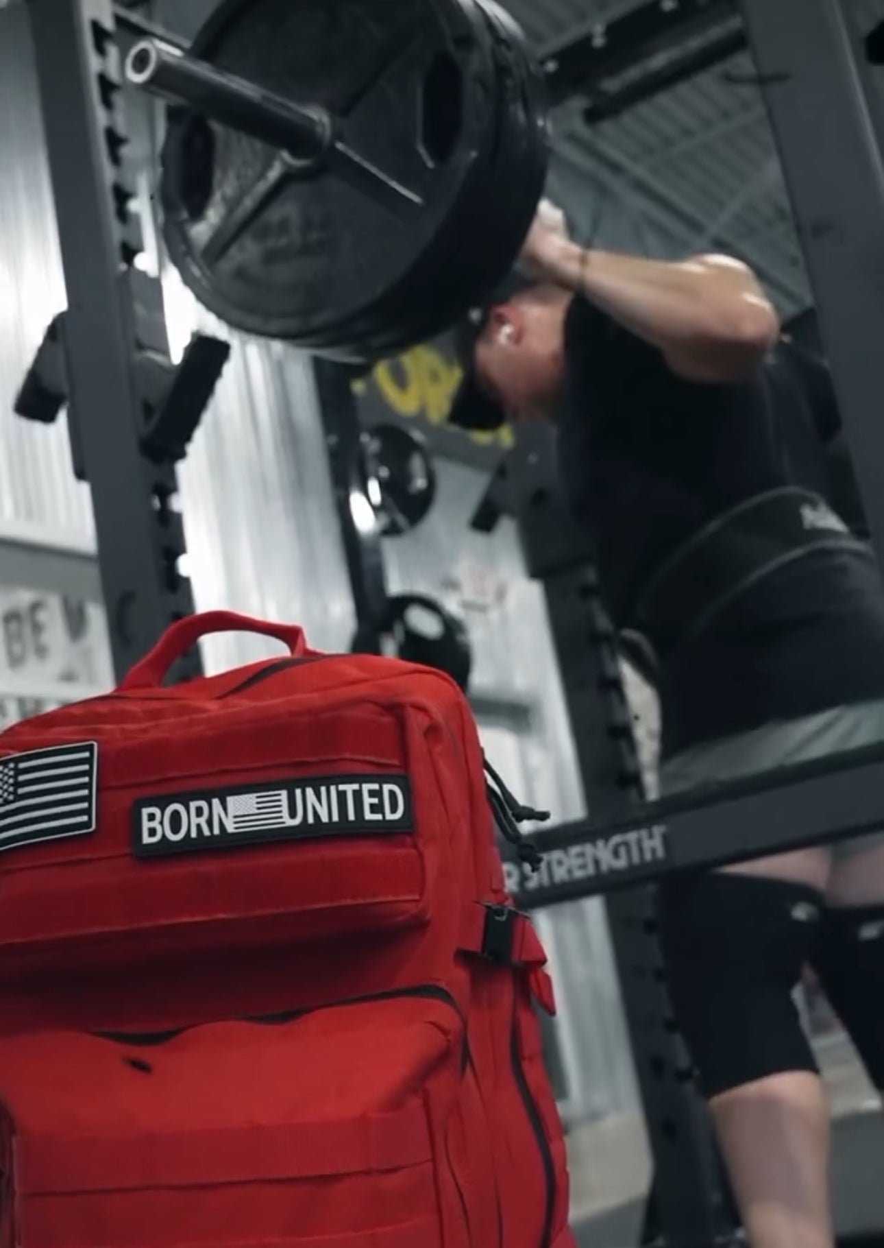 In a gym, someone squats with heavy weights on a barbell. In the foreground, a Revenge Red Tac-Pac - 45L by Born United Private Label with an American flag patch stands out against metal walls and strength equipment, embodying determination and resilience.
