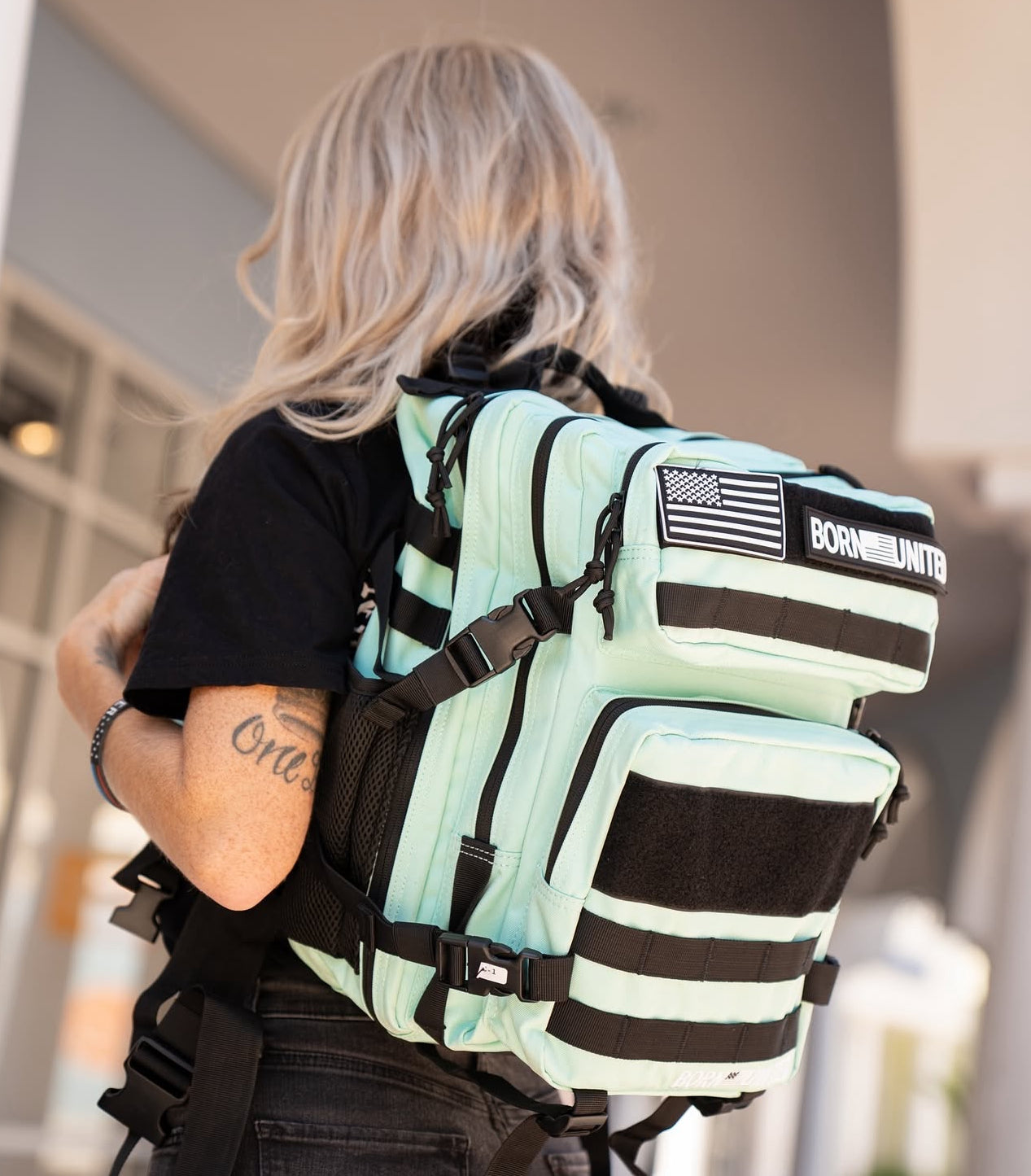 A person with long blonde hair sports a Born United Private Label Mint Condition Premium Tac-Pac - 25L in mint green with black straps, featuring a USA flag patch and BORN UNITED text. Outdoors, they proudly display their tattooed arm.