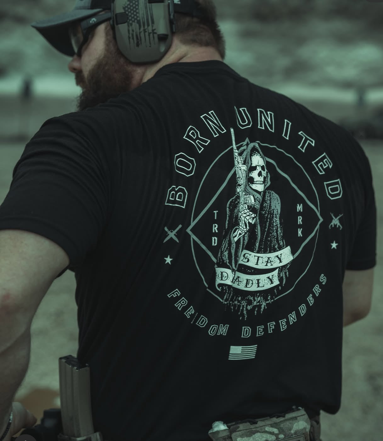 Wearing the Stay Deadly Tee - Black by Born United Private Label, featuring a Grim Reaper with a rifle and the text BORN UNITED, STAY DEADLY, and FREEDOM DEFENDERS, paired with headphones and camo pants for a stylish, fierce look.
