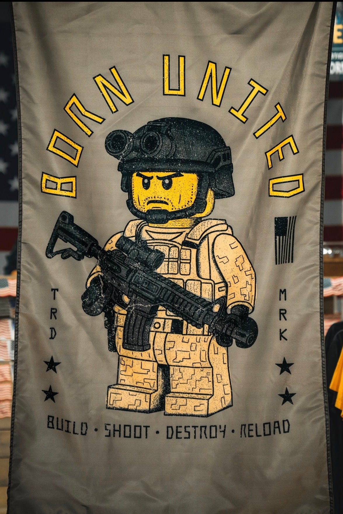 The Brick Operator Flag by Born United Private Label features a LEGO-style soldier in desert camo with a rifle, set against an American flag and surrounded by “BORN UNITED,” “BUILD SHOOT DESTROY RELOAD,” and stars and stripes.