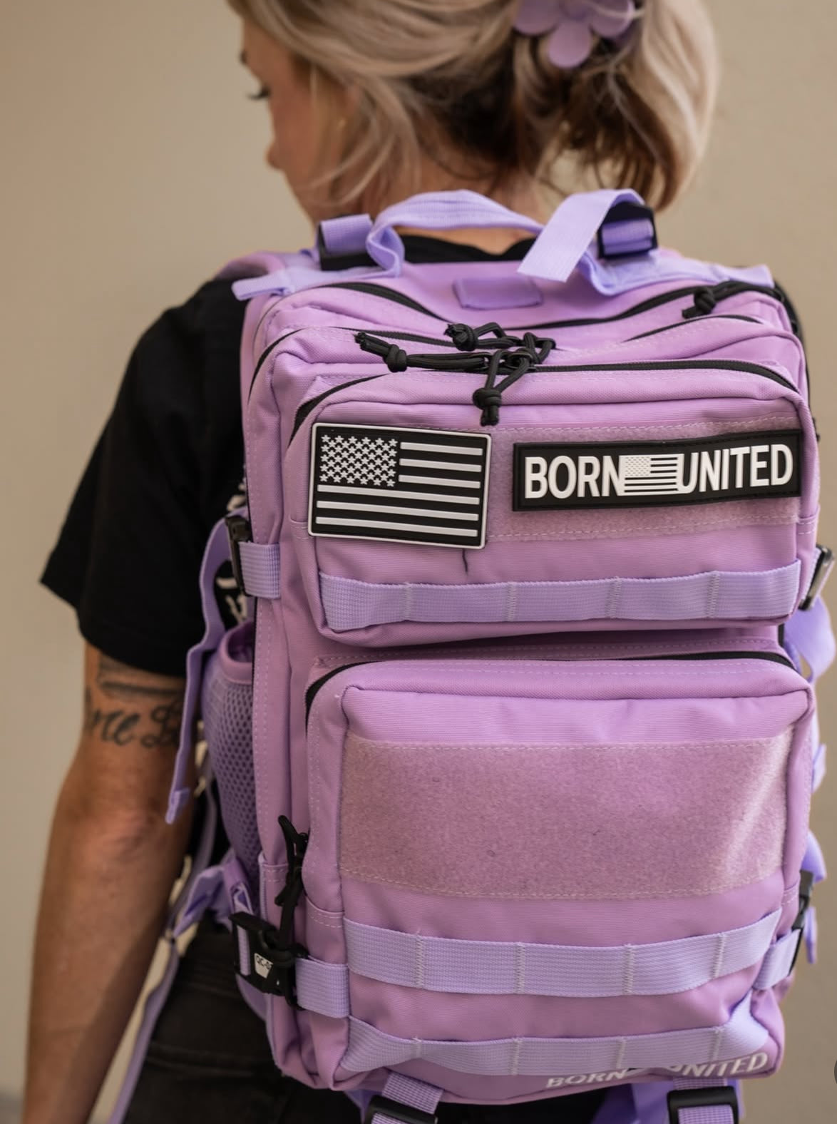 A blonde-haired person is sporting the Imperial Purple Tac-Pac - 25L from Born United Private Label. It includes black-and-white patches reading BORN UNITED with an American flag and offers multiple compartments and adjustable straps for convenience.