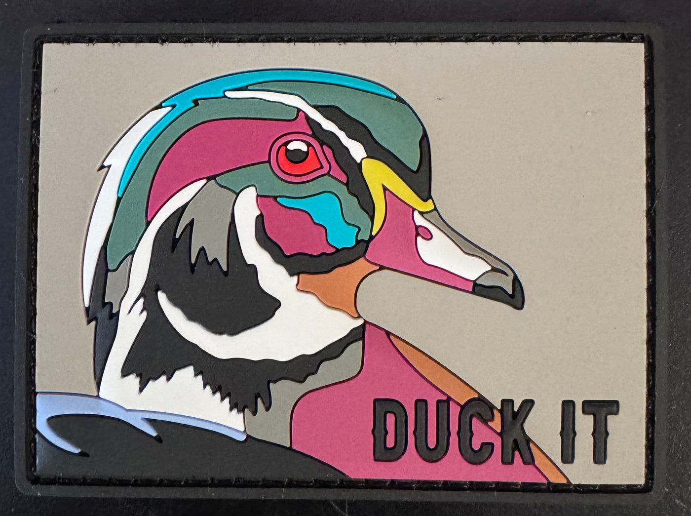 A colorful ducks head with vibrant patterns on a gray background is featured on the Mucker Buckets Duck It PVC Patch, with the phrase boldly written in black at the bottom right.