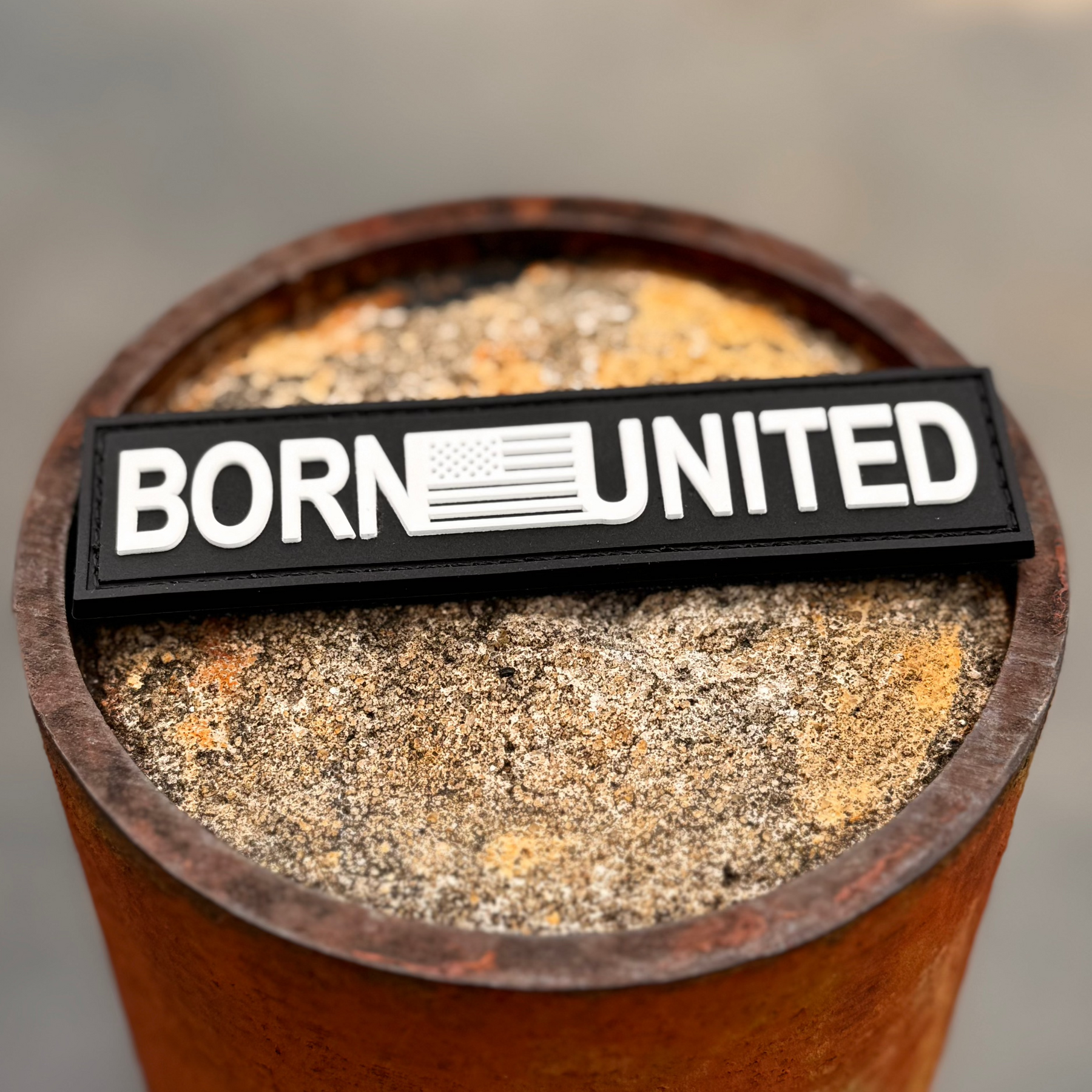 The Born United Private Label Born United PVC Patch, featuring a US flag as the O, rests on a rusted metal cylinder filled with sand or concrete—ideal for Tac-Pacs fans.