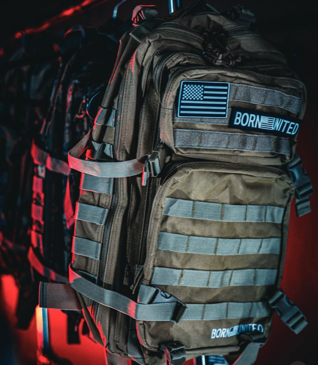 The Coyote Brown Tac-Pac - 45L by Born United Private Label, complete with an American flag and BORN UNITED patches, is displayed. The backdrop of similarly styled bags under red lighting adds intrigue to the scene.