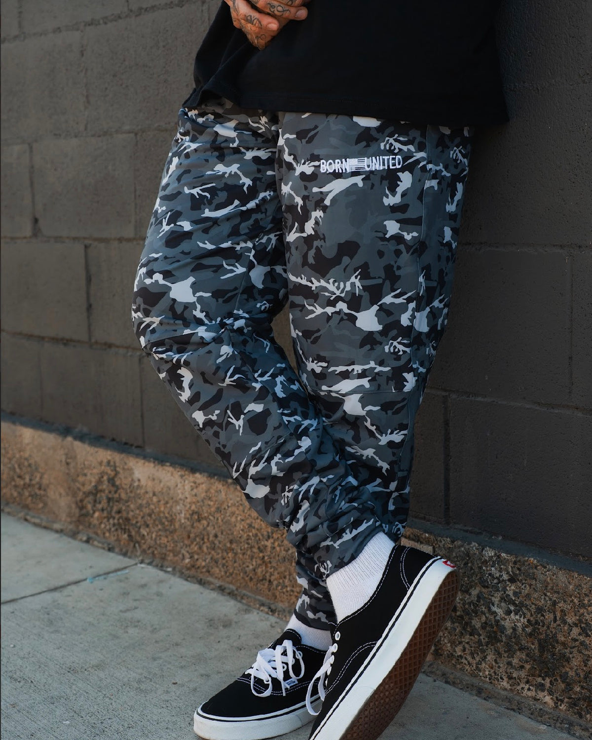 A person stands against a dark brick wall with one foot crossed, wearing Born United Private Label Men's Classic Premium Joggers in Gunmetal Camo, black Vans sneakers with white laces, and white socks.