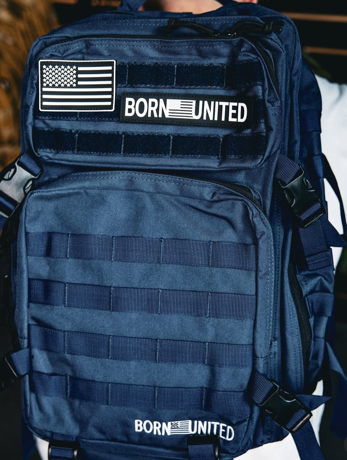 The Navy Blue Tac-Pac - 45L by Born United Private Label showcases the BORN UNITED logo and an American flag patch on the front. With multiple straps and zippers, this durable, functional design is ideal for outdoor or military use.
