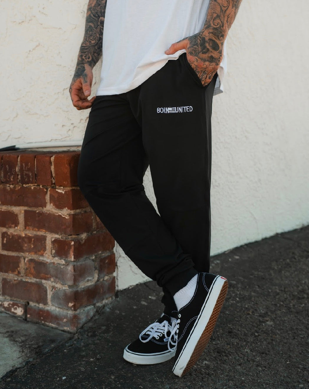 A person with tattooed arms, dressed in a white t-shirt, black Vans, and Born United Private Label Men's Classic Premium Joggers | Stealth, stands casually against a brick ledge and white wall.