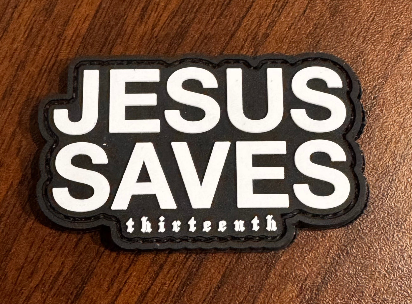 A black Jesus Saves PVC Patch by 13th features bold white JESUS SAVES text and smaller thirteenth below, shown on a wooden surface.
