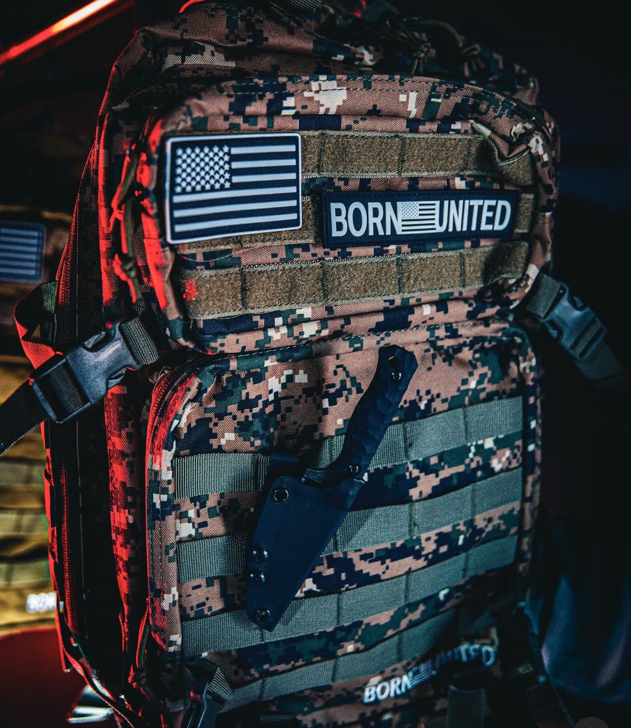 The Digi-Cam(Bechtold) Tac-Pac - 45L by Born United Private Label boasts an American flag patch and BORN UNITED text. A sheathed knife is attached at the front. Dim lighting with red highlights complements its rugged, tactical look, perfect for adventurous digital camera photographers.