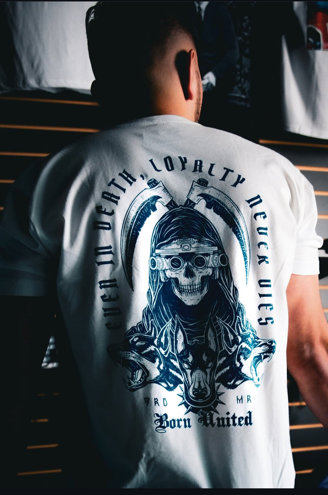 A person wears a white tee from Born United Private Labels Loyalty line, featuring a skeleton in a hood and goggles with two wolves and the text, Even in death, loyalty never dies, and Born United.