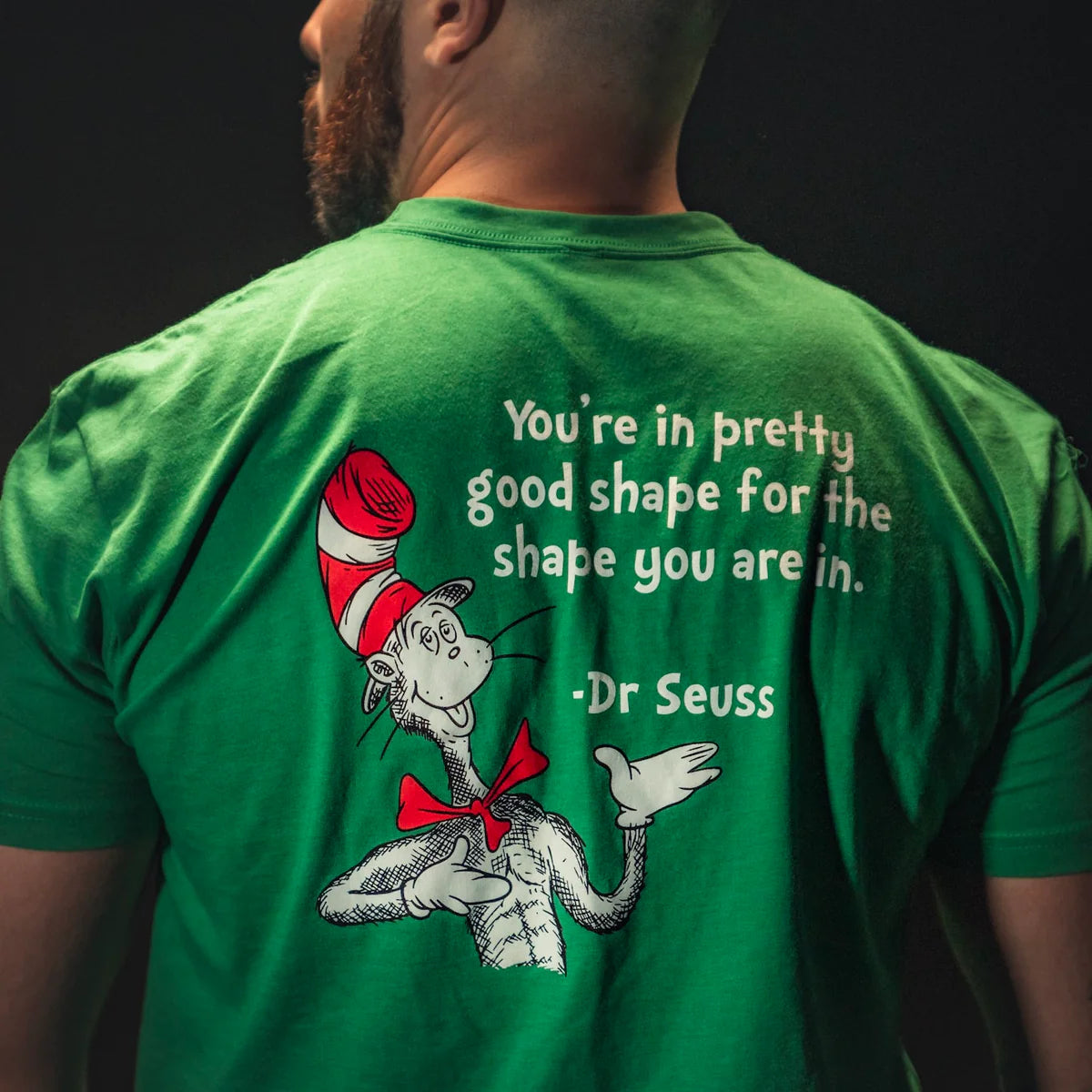 A man wears the St. Seuss Tee from Tango Charlie Apparel, featuring a green design with a Dr. Seuss character in a red hat and bow tie, along with the text: Youre in pretty good shape for the shape you are in, symbolizing resilience and self-improvement through whimsical wisdom.