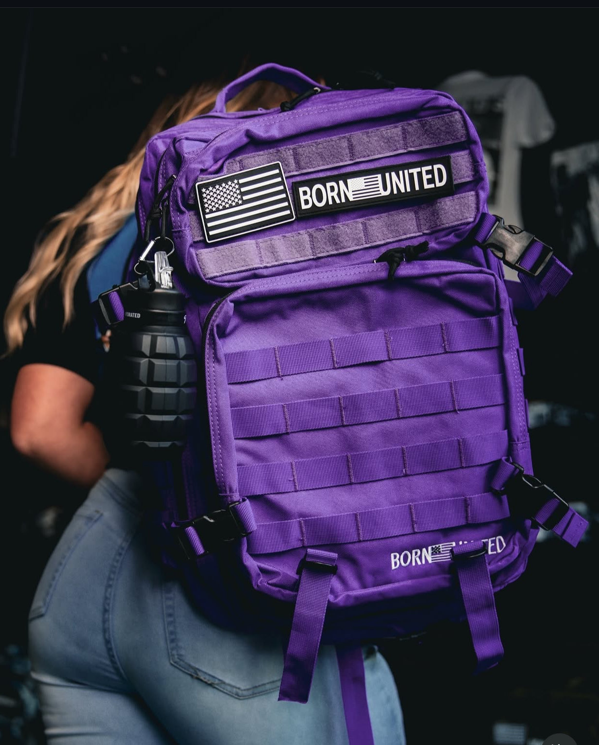 A person carries a Cosmic Purple Tac-Pac - 45L by Born United Private Label, complete with American flag patches and BORN UNITED text. A black grenade-style accessory on the side perfectly complements their blue jeans.