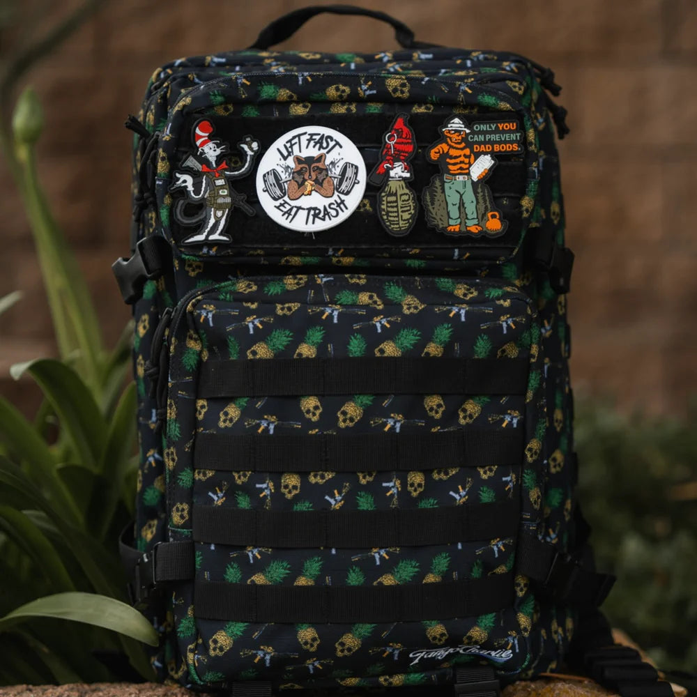The TCA Tac-Pac Tropic Thunder 2.0 by Born United Private Label is a black backpack with colorful skull and cartoon patches, offering tactical versatility. With its waterproof design, its perfect for outdoor adventures and stands out in lush green surroundings.